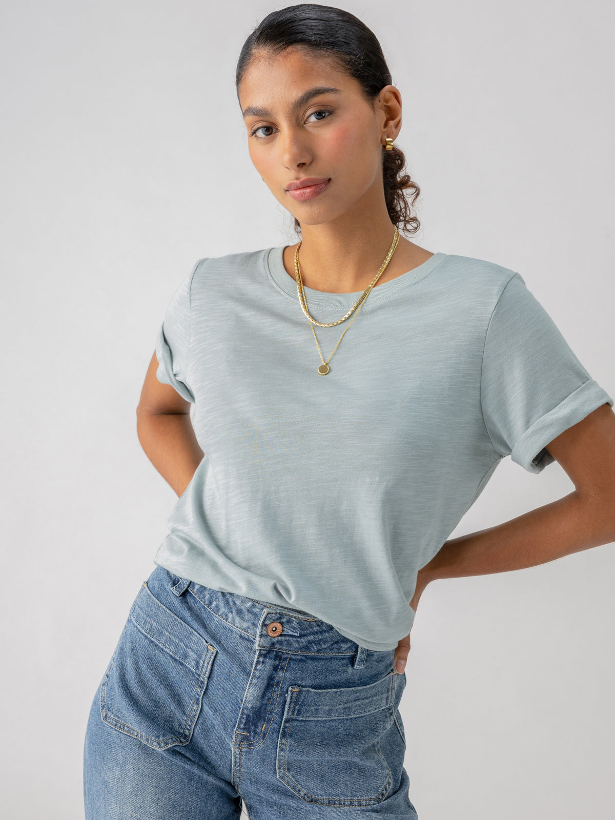 A person with dark pulled-back hair stands confidently. They wear the Perfect Tee Remix Eucalyptus from Sanctuary Clothing, light blue jeans, and layered gold necklaces. The neutral background enhances the focus on their casual yet stylish outfit.