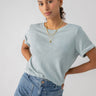 A person with dark pulled-back hair stands confidently. They wear the Perfect Tee Remix Eucalyptus from Sanctuary Clothing, light blue jeans, and layered gold necklaces. The neutral background enhances the focus on their casual yet stylish outfit.