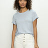 A person with curly hair poses against a neutral backdrop, wearing the perfect tee remix sky blue by Sanctuary Clothing and high-waisted white pants with buttons. They have a calm expression, hands relaxed at their sides.