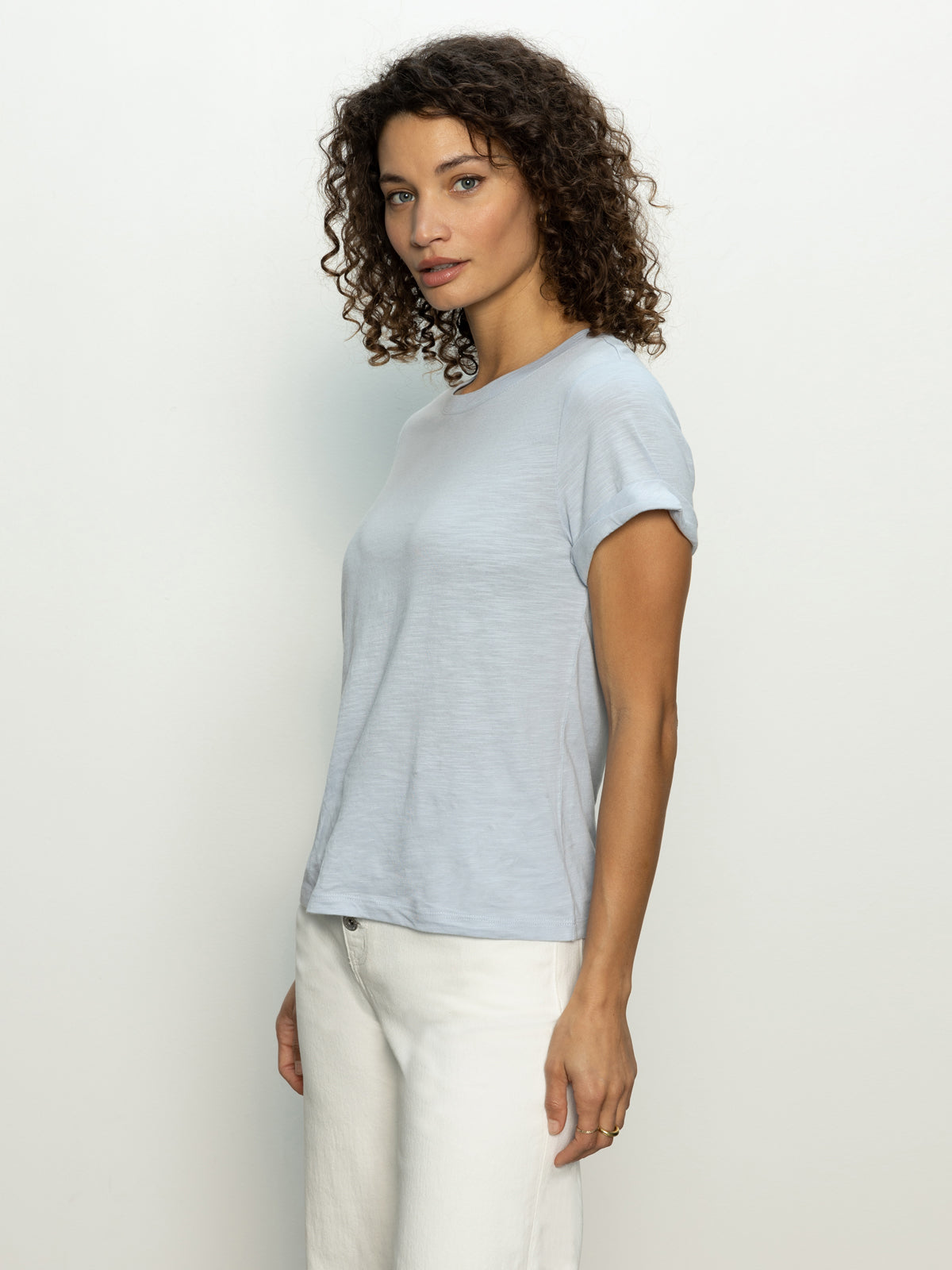 A person with curly hair stands against a plain background, facing slightly to the side. They wear a sky blue Perfect Tee Remix by Sanctuary Clothing and white pants, with a calm expression.