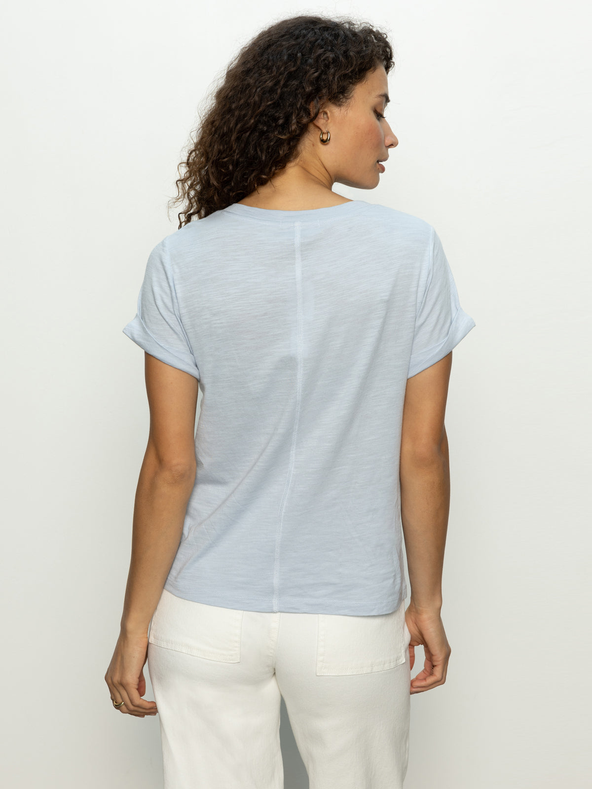 A person with curly hair, facing away, wears the "Perfect Tee Remix" in sky blue by Sanctuary Clothing and white pants. The shirt features rolled sleeves and a seam down the back against a plain white background.