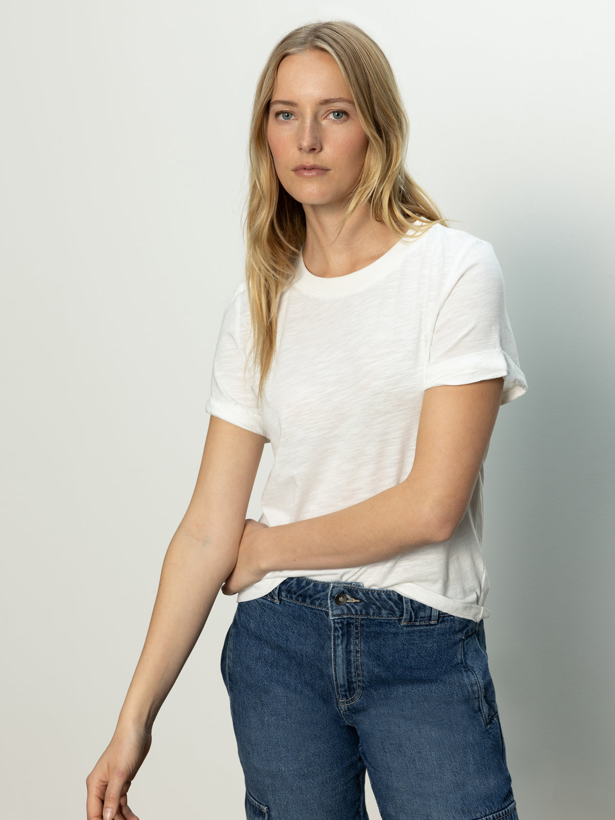 A woman with long blonde hair stands against a plain background, wearing Sanctuary Clothing's Perfect Tee Remix in white and blue jeans. Her expression is neutral, with her left arm relaxed by her side.