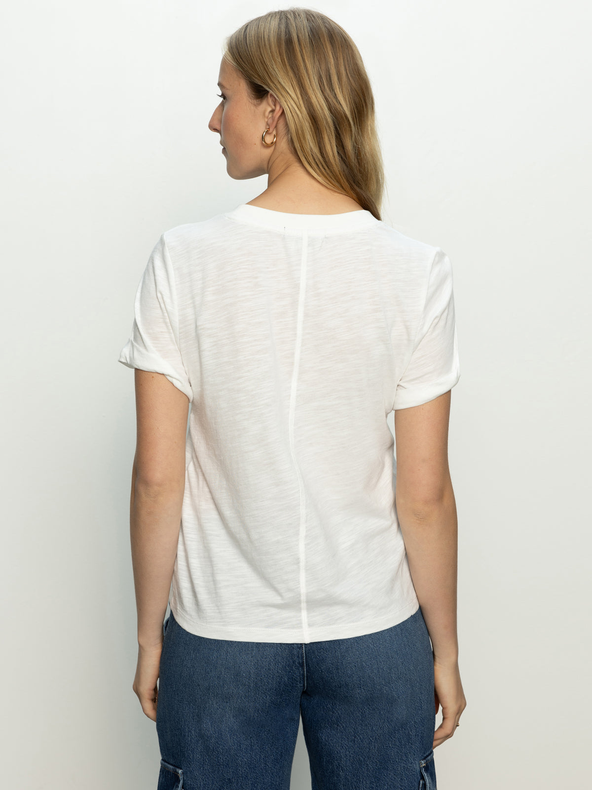 A person with long hair is standing with their back to the camera, wearing Sanctuary Clothing's perfect tee remix in white and blue jeans. The background is a plain off-white.