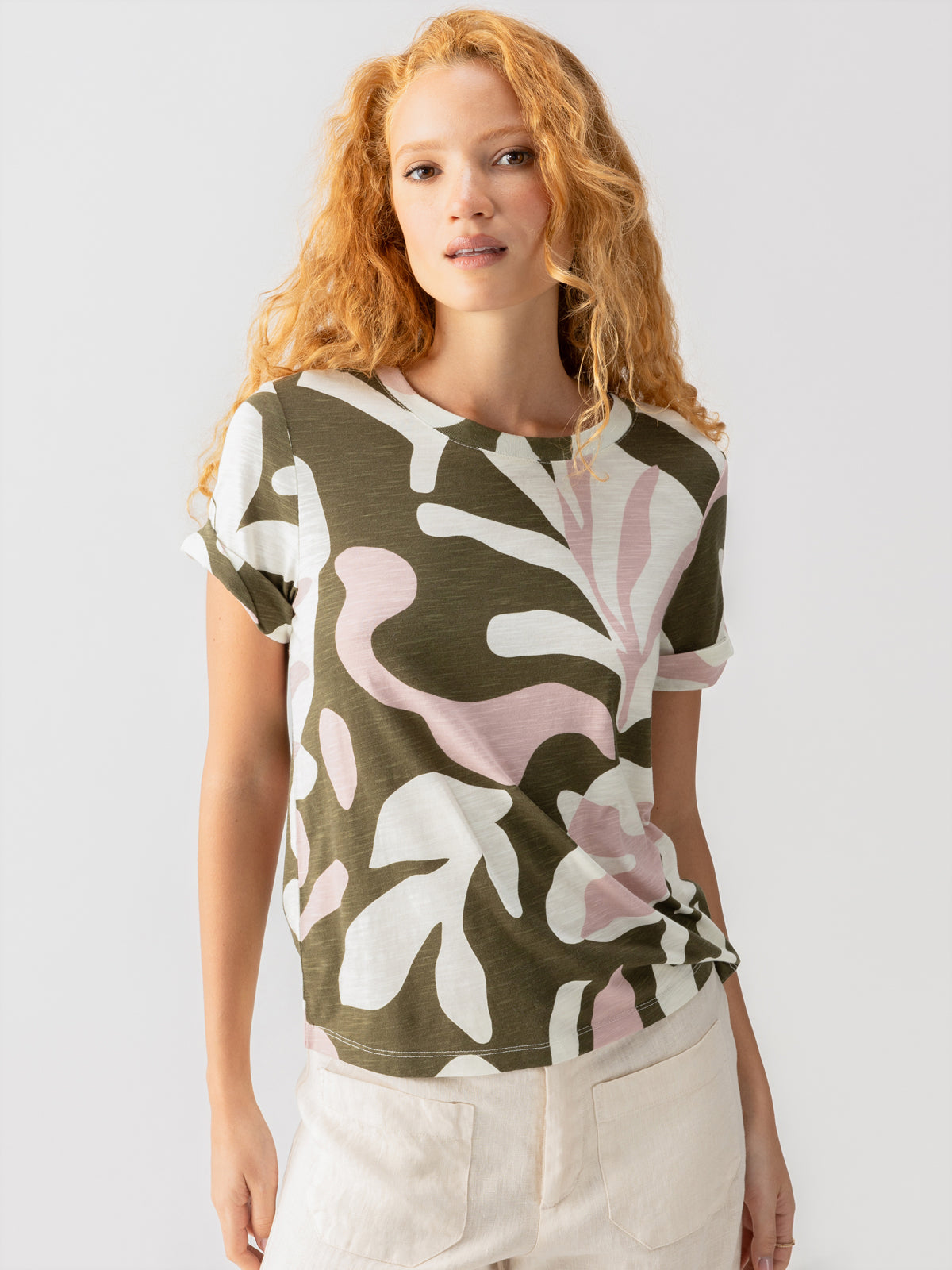 A person with long, wavy red hair is wearing the Perfect Tee Remix Fatigue Mineral from Sanctuary Clothing, featuring a green, white, and pink abstract leaf pattern, paired with light-colored shorts. The individual is standing against a plain white background, looking slightly to the side with a neutral expression.