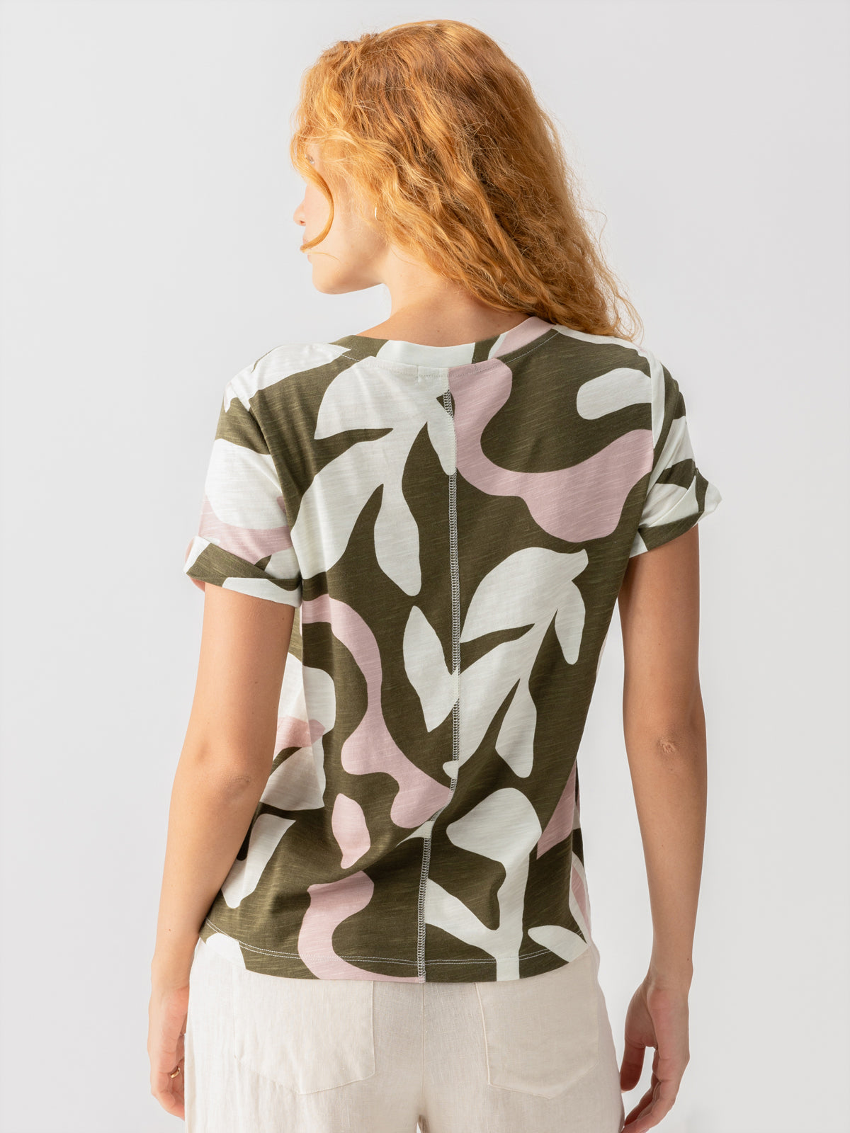 A woman with long wavy red hair stands facing away, wearing the "Perfect Tee Remix Fatigue Mineral" by Sanctuary Clothing, showcasing its short sleeves and camouflage pattern in shades of green, white, and pink. She pairs it with light-colored pants against a plain, light-colored background.