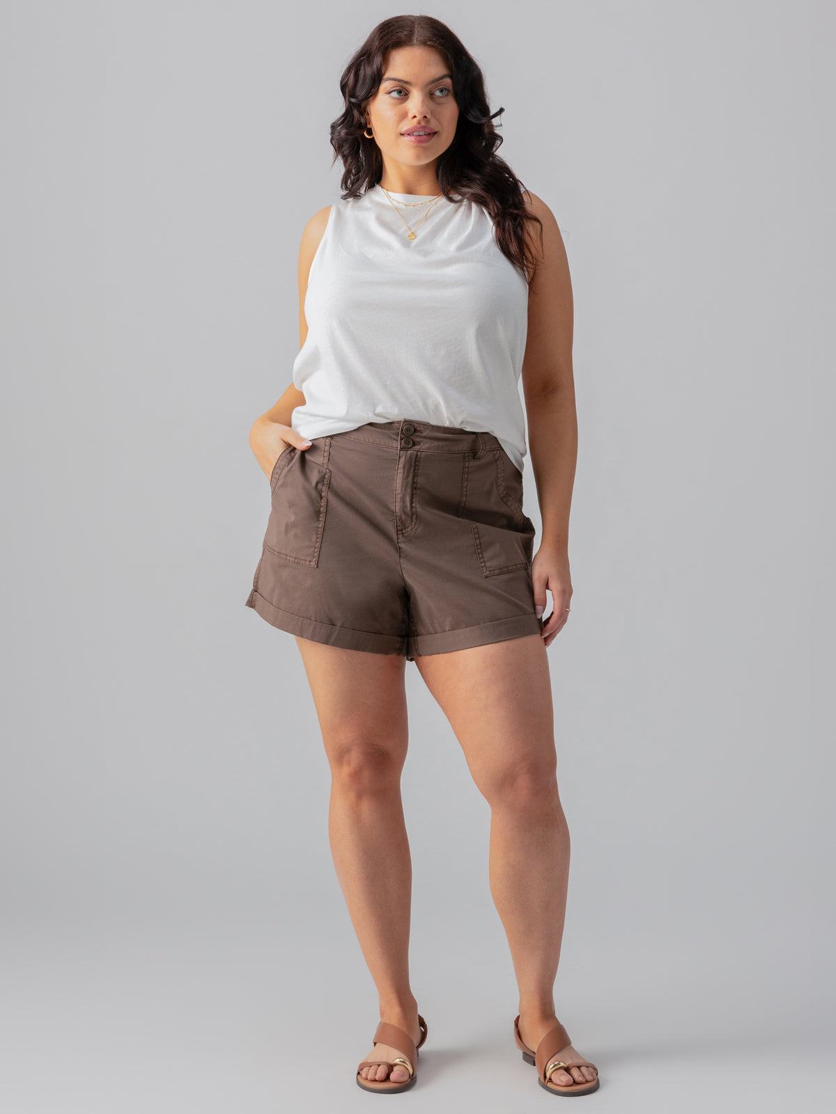 A woman stands in front of a plain backdrop, wearing the "Suns Out Tee White" from the Sanctuary Clothing Inclusive Collection and brown high-waisted shorts. She has long, dark, wavy hair and is wearing brown sandals. Her left hand is in her pocket, and she has a slight smile. She accessorizes with a necklace.