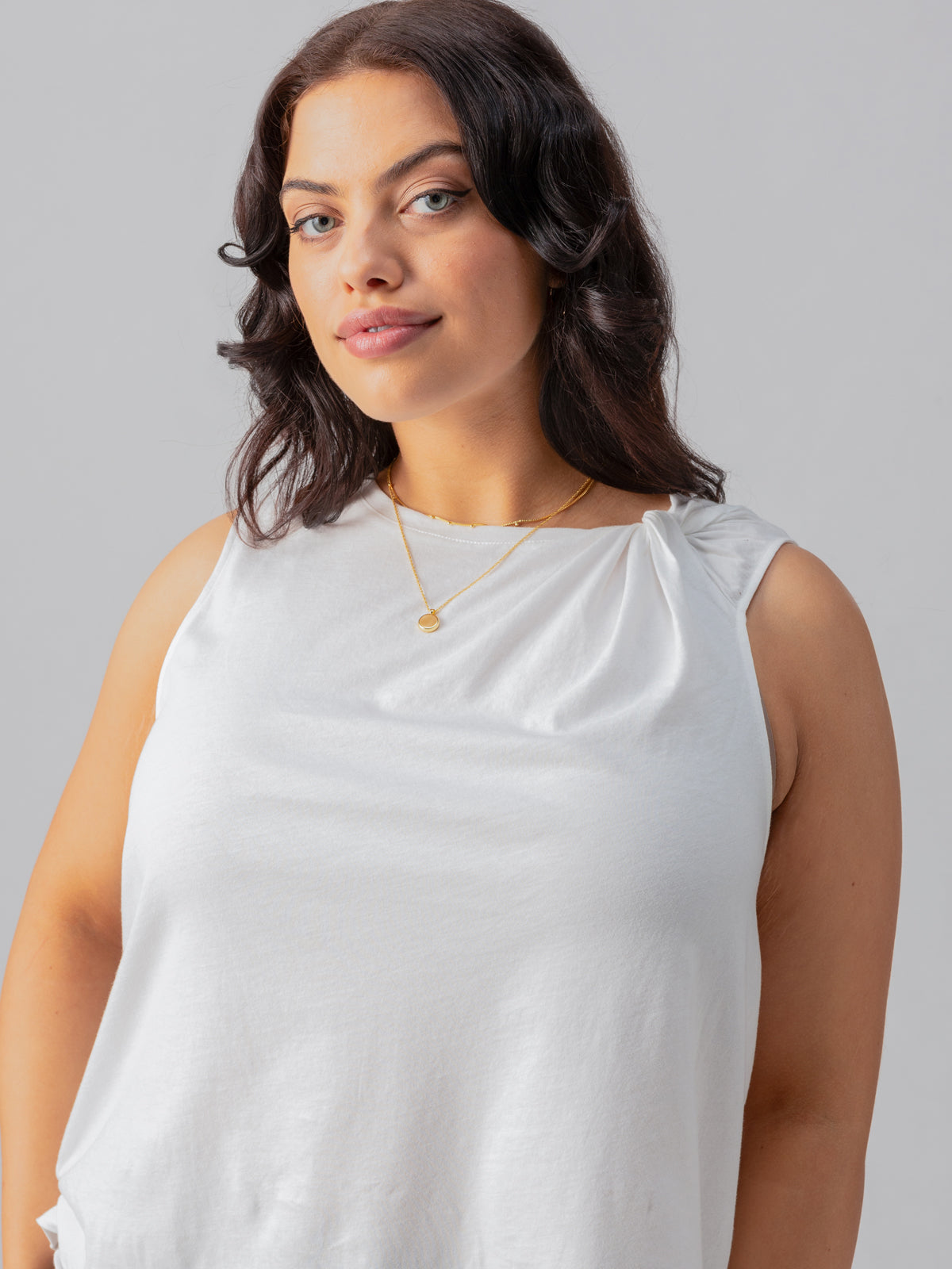 A woman with dark wavy hair, wearing the "Suns Out Tee" from Sanctuary Clothing's Inclusive Collection and a gold necklace, stands against a gray background. She has a slight smile and looks confidently at the camera.