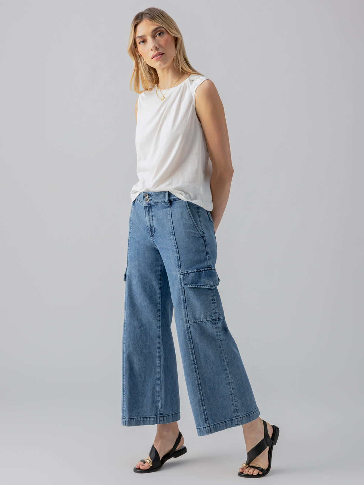 A person with long, light-colored hair is donning the Suns Out Tee White by Sanctuary Clothing, paired with wide-legged blue denim jeans featuring pockets. They are standing in a relaxed pose, hands in pockets, and wearing black sandals. The backdrop is plain and light-colored.