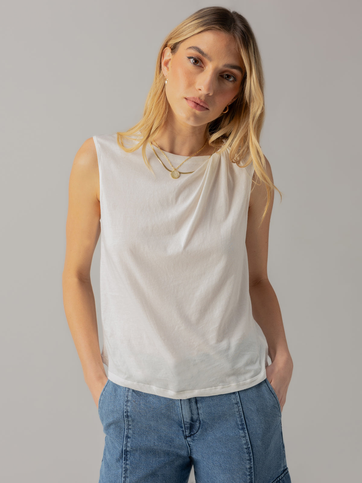 A person with long, blonde hair wearing a Suns Out Tee White by Sanctuary Clothing and blue jeans stands against a neutral background. They are gazing slightly to the side with hands in their pockets, wearing two layered necklaces.