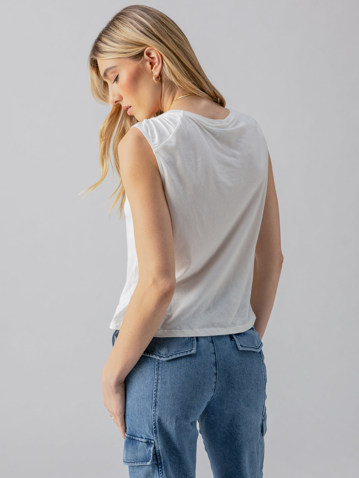 A person with long blonde hair is seen from behind, wearing a sleeveless white "Suns Out Tee" by Sanctuary Clothing and blue jeans. The background is plain and light-colored, emphasizing the casual outfit.