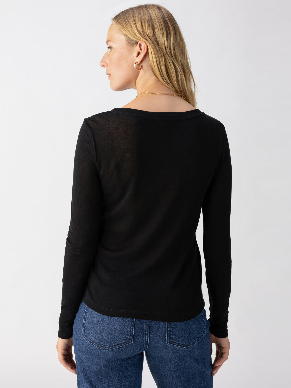 A person with long blonde hair is standing with their back to the camera. They are wearing a long-sleeved Femme Crew Tee in black by Sanctuary Clothing and blue jeans. The background is plain and light in color.