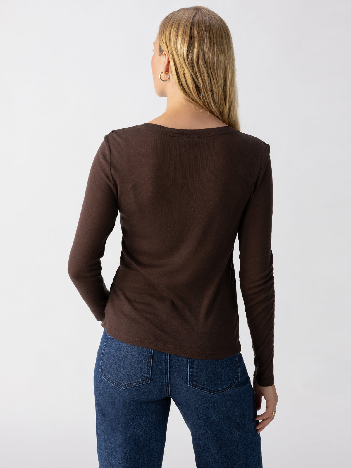 A person with long blonde hair is standing with their back to the camera. They are wearing a Sanctuary Clothing Femme Crew Tee Coffee and blue jeans. The background is plain and light-colored.