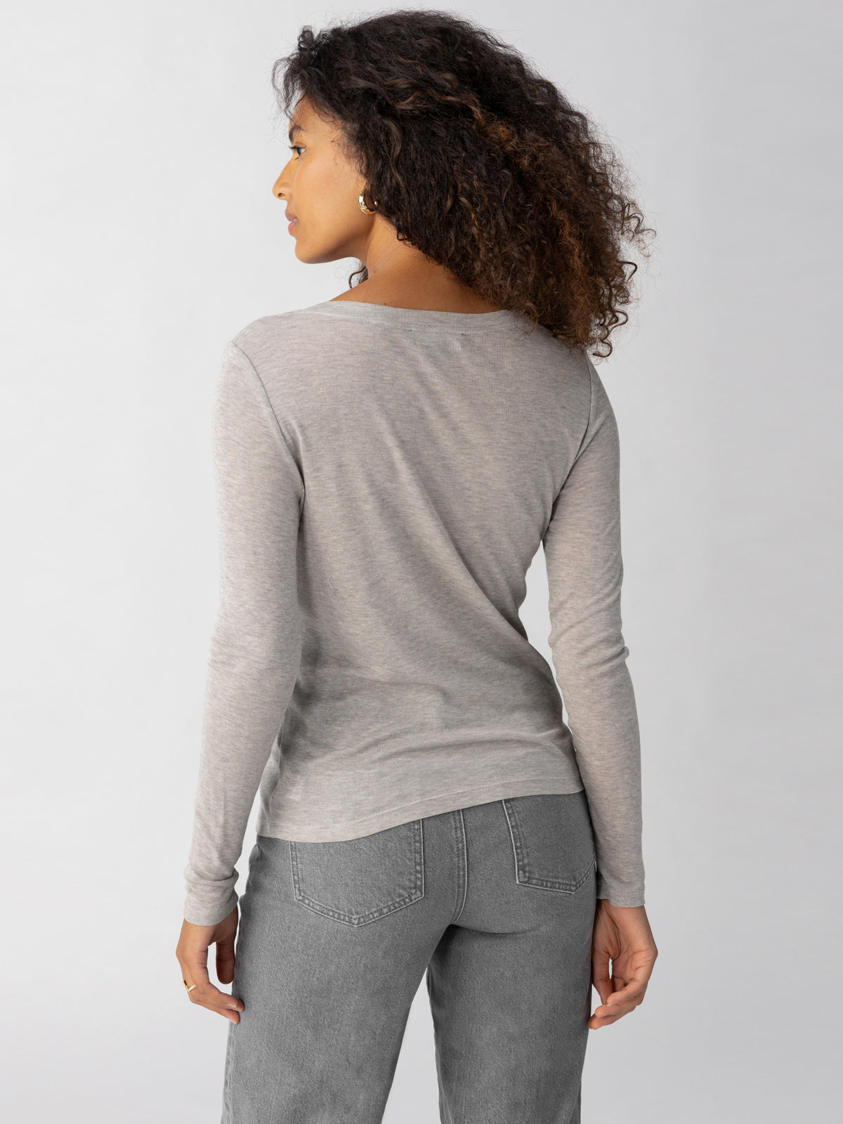 A person with curly hair, wearing the Sanctuary Clothing Femme Crew Tee Heather Grey and gray pants, is standing and facing away from the camera, showing a side profile. The background is plain and light-colored.