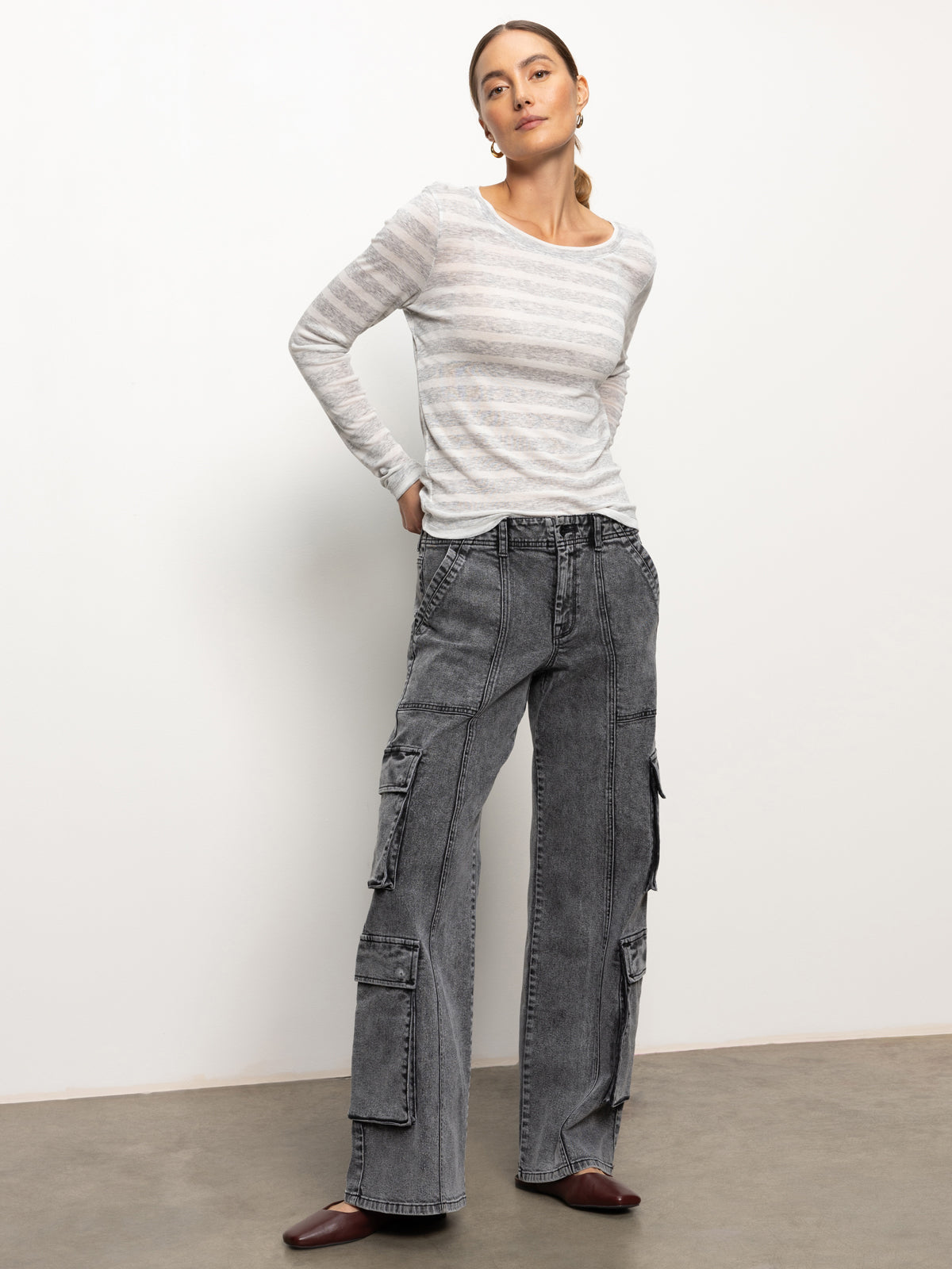 A person stands against a plain background, wearing the femme crew stripe tee dewey grey chalk stripe from Sanctuary Clothing, and gray cargo pants with multiple pockets. They have their hands in their pockets and are wearing brown shoes.