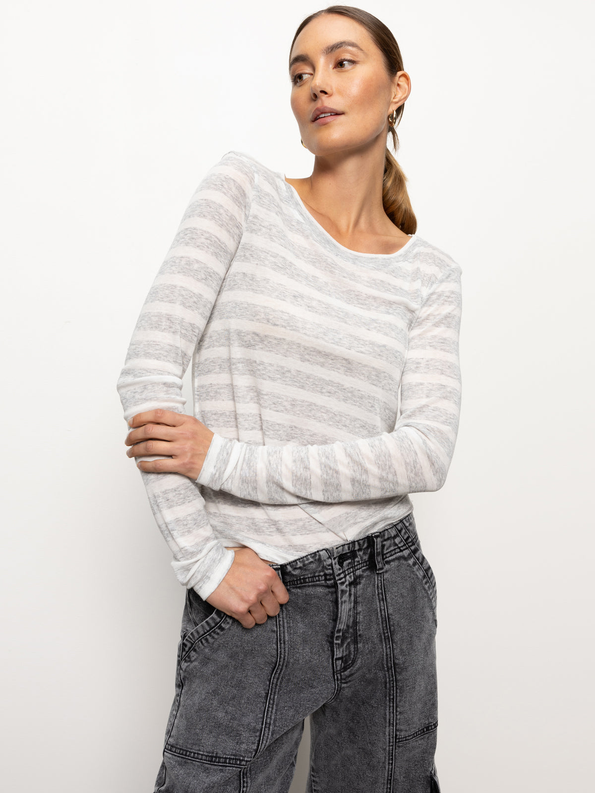 A person dressed in the femme crew stripe tee dewey grey chalk stripe by Sanctuary Clothing and dark gray jeans is standing against a plain background. Their light brown hair is pulled back, and they're looking upward with a slight smile, arms crossed.