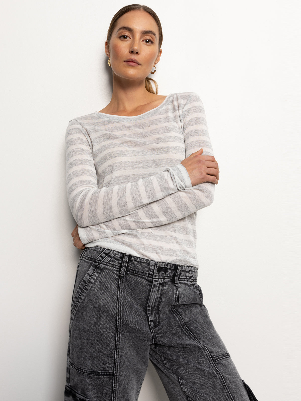 A person with long hair is standing against a white background, wearing the femme crew stripe tee by Sanctuary Clothing in dewey grey chalk stripe along with gray denim pants. They have their arms crossed and are looking confidently at the camera.