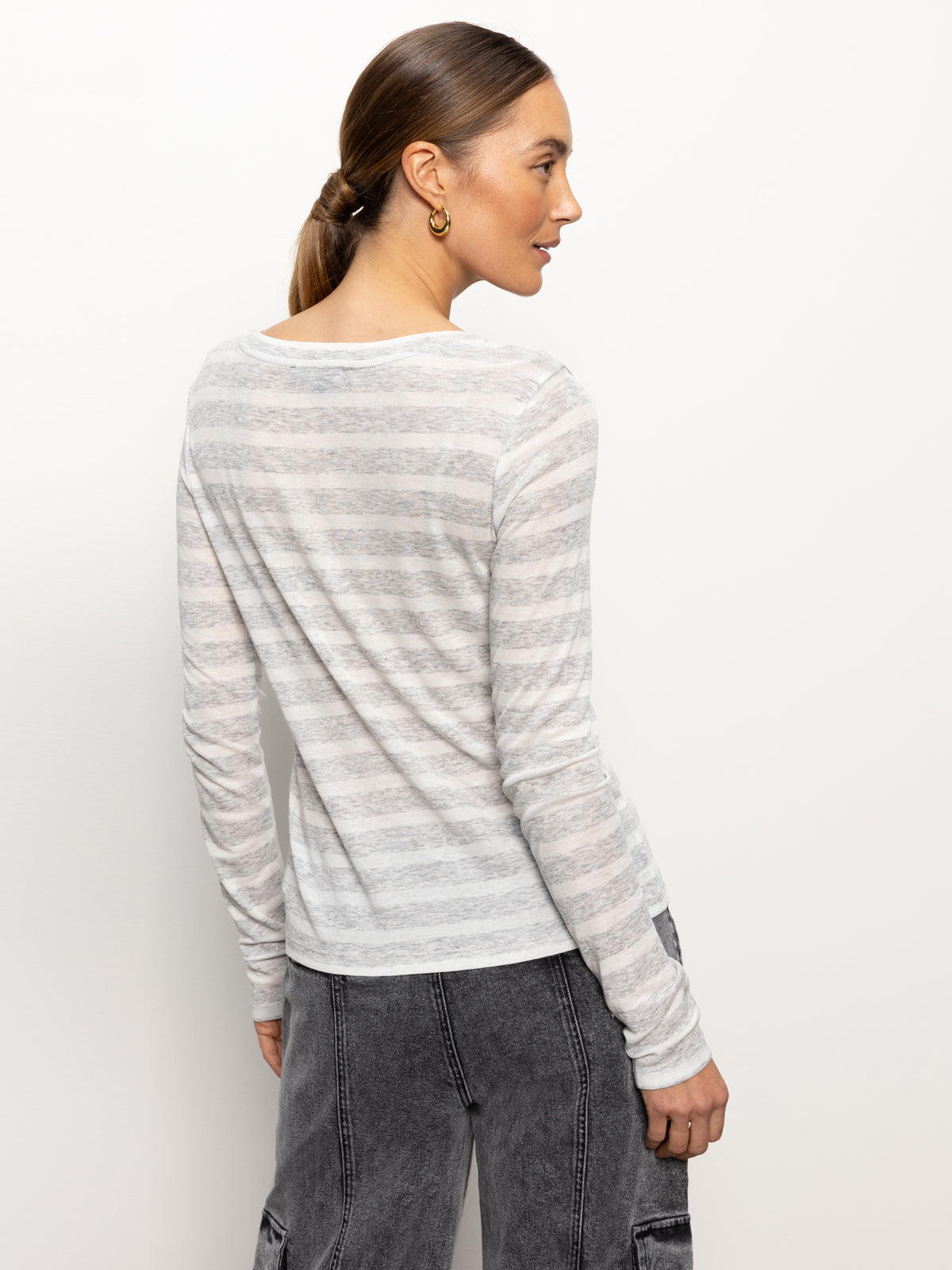 A person with long hair in a ponytail is wearing the "femme crew stripe tee dewey grey chalk stripe" by Sanctuary Clothing, paired with dark gray cargo pants. They are standing against a plain white background, facing to the side.