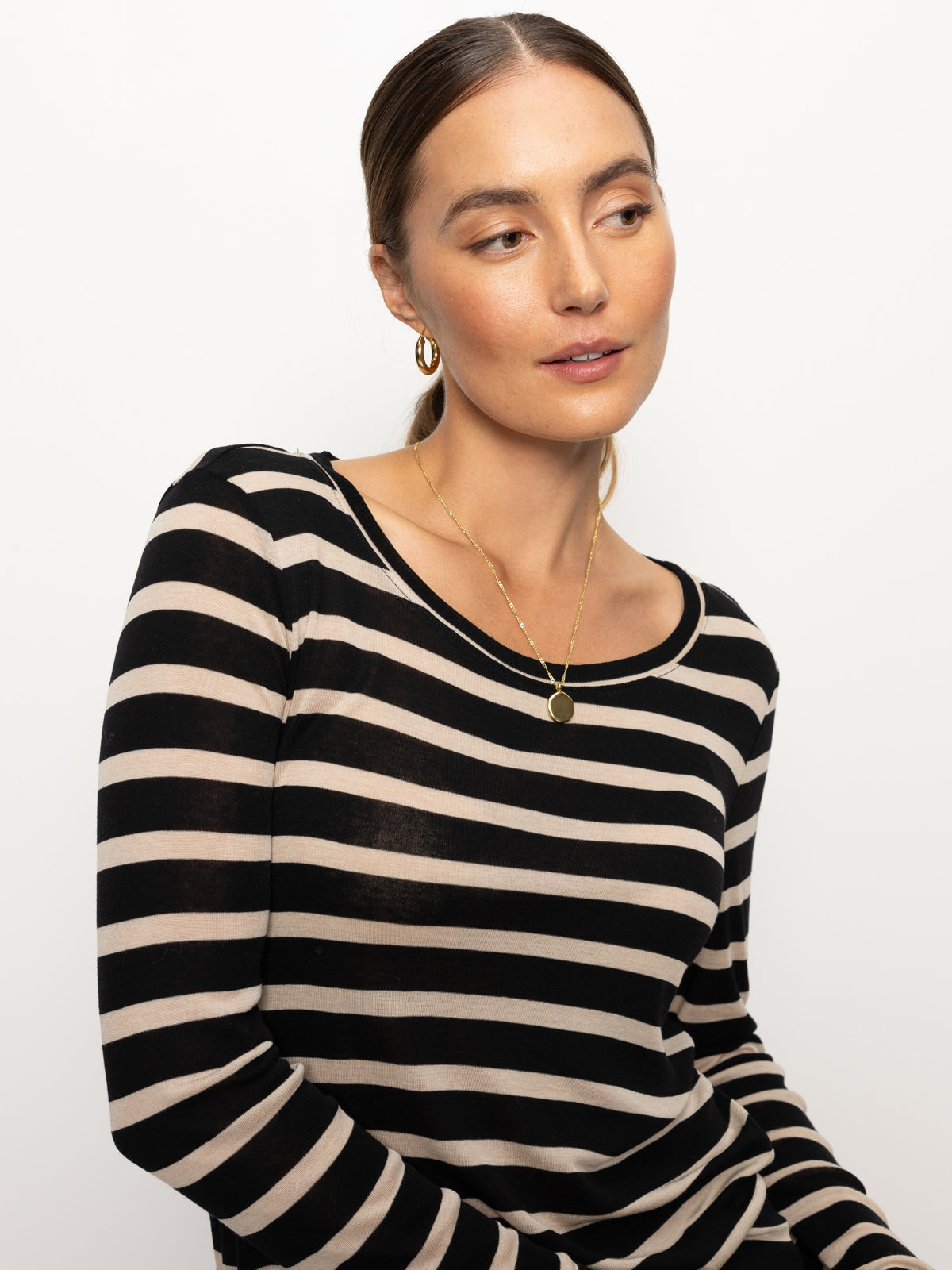 A person with long hair is wearing the femme crew stripe tee in oat black stripe by Sanctuary Clothing and gold hoop earrings. They are looking to the side with a neutral expression, set against a plain white background.