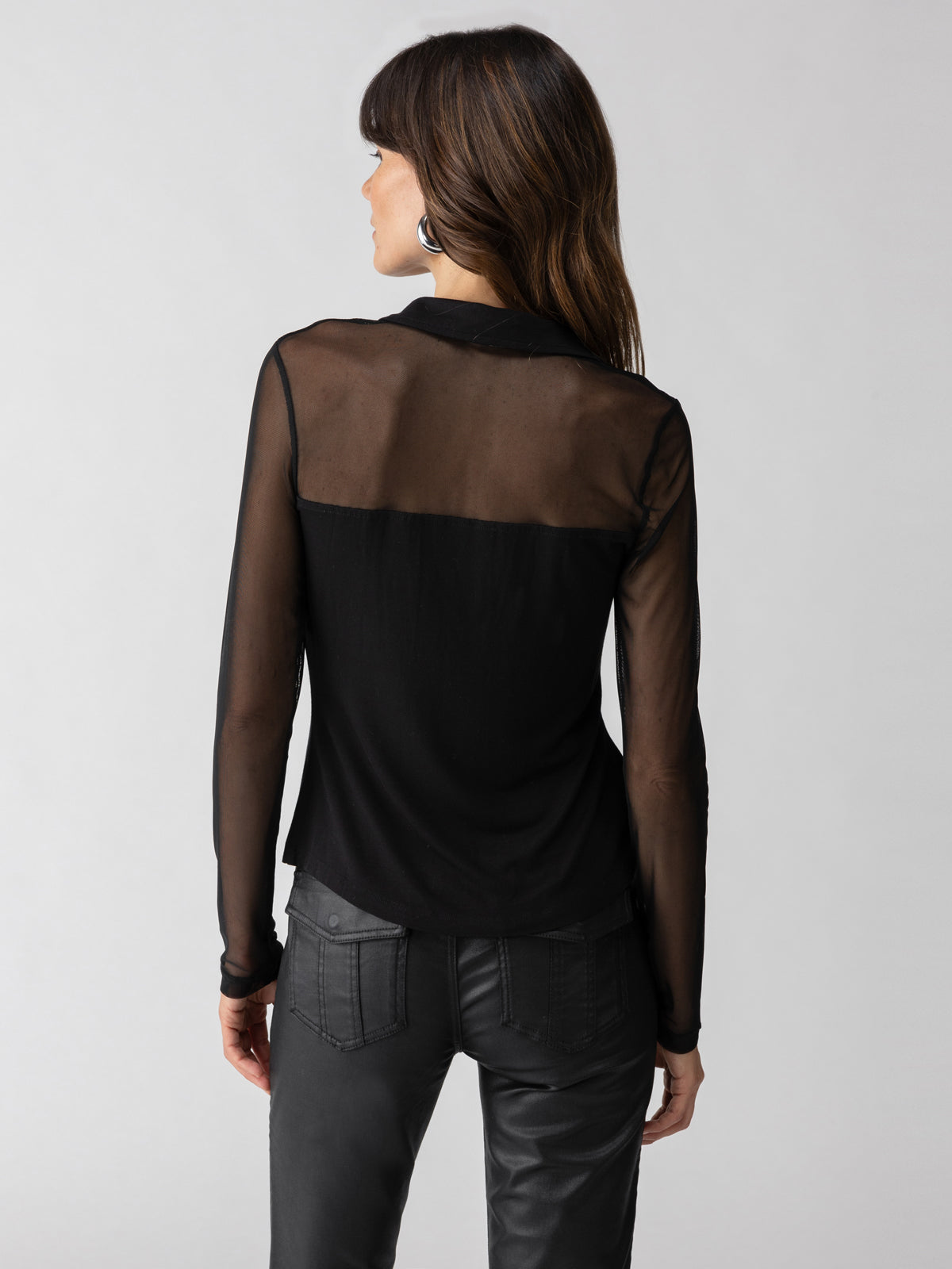 A woman with long brown hair is facing away from the camera. She is wearing the "Take Me Away Top Black" by Sanctuary Clothing, a sheer black blouse with a solid back yoke and long sleeves, paired with black pants. The background is a plain, light gray color.