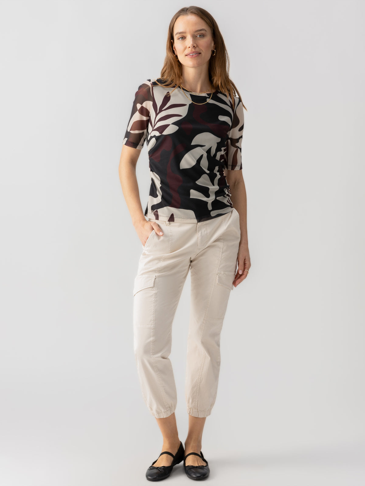 A woman with long hair is standing against a plain backdrop. She is wearing the Half Sleeve Perfect Mesh Tee Mineral from Sanctuary Clothing, which features a black and white abstract pattern, paired with white cargo pants with side pockets and black flats. She has a relaxed expression with one hand in her pocket.