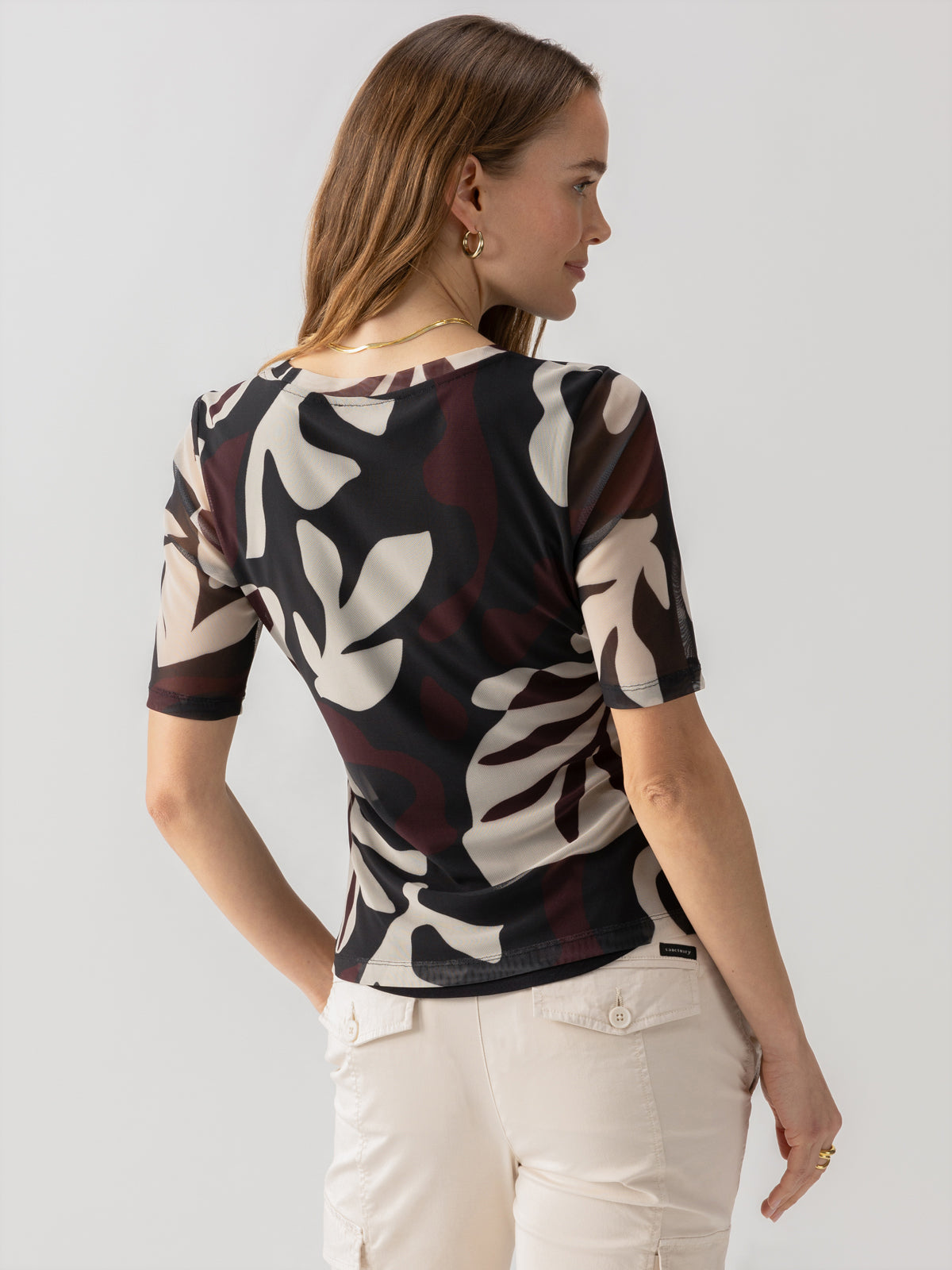 A woman with long, light brown hair is seen from the back. She is wearing the Half Sleeve Perfect Mesh Tee Mineral by Sanctuary Clothing, which features a black, gray, cream, and maroon abstract pattern. She has paired it with light-colored pants that have pockets. She is looking slightly to her right.