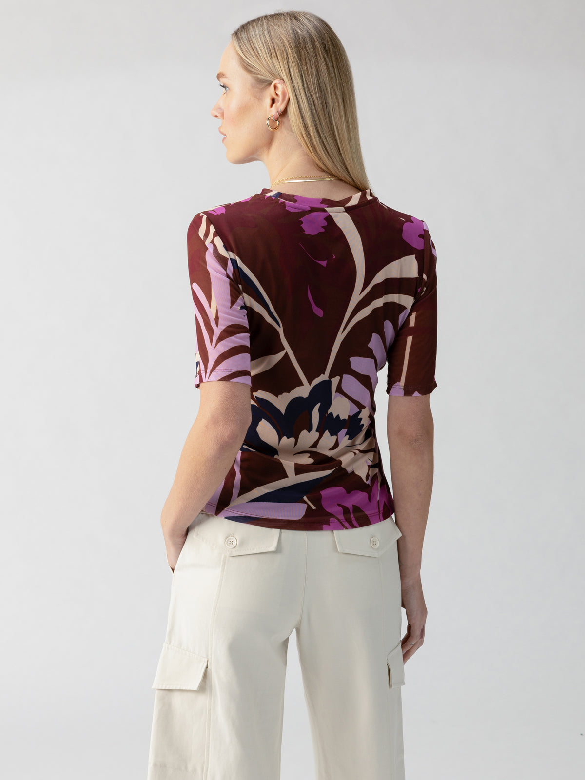 A person with long blonde hair is seen from the back, wearing the Half Sleeve Perfect Mesh Tee in South Palm Rich Clay by Sanctuary Clothing and light-colored cargo pants. The person has their hands in their pockets and is standing against a simple, light-colored background.