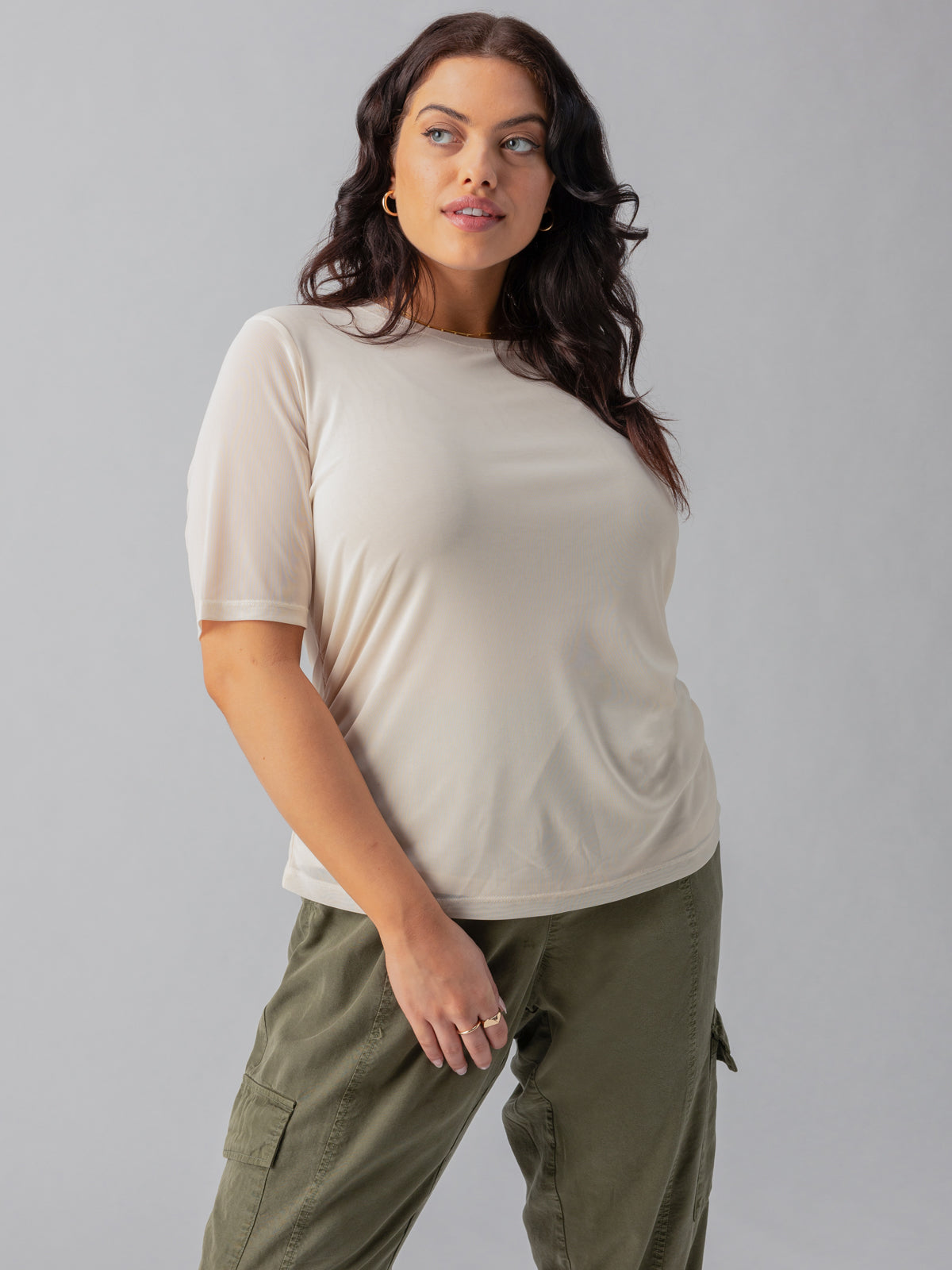 A woman with wavy dark hair is wearing a Sanctuary Clothing Half Sleeve Perfect Mesh Tee Birch from the Inclusive Collection and green cargo pants. She stands against a plain gray background, looking confidently towards the camera with a slight smile on her face, her left hand resting on her hip.