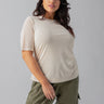 A woman with wavy dark hair is wearing a Sanctuary Clothing Half Sleeve Perfect Mesh Tee Birch from the Inclusive Collection and green cargo pants. She stands against a plain gray background, looking confidently towards the camera with a slight smile on her face, her left hand resting on her hip.