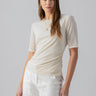 A woman with long brown hair stands against a plain background, wearing the fitted Sanctuary Clothing Half Sleeve Perfect Mesh Tee in Birch, paired with white pants featuring side pockets. She accessorizes with a gold necklace and hoop earrings. Her expression is neutral, with a slight tilt to her head.