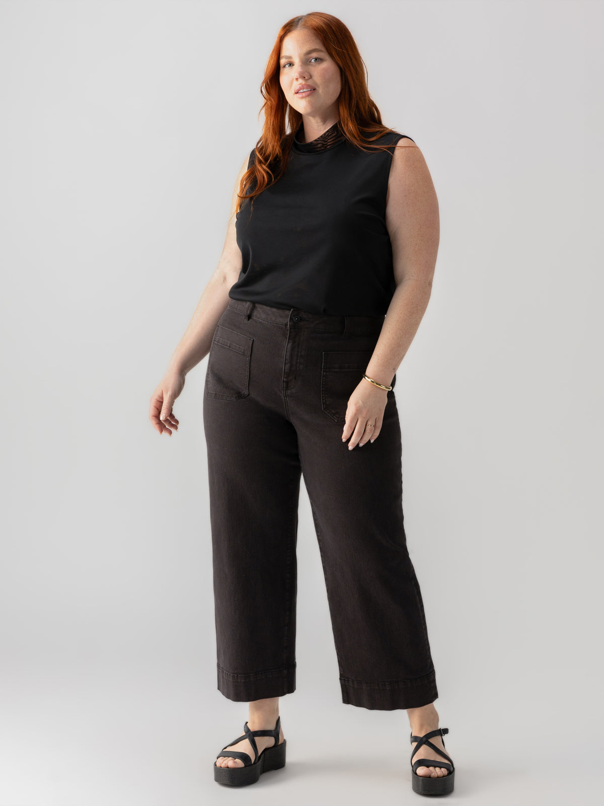 A woman with long red hair is standing against a plain white background, confidently gazing at the camera. She is dressed in the Shirred Neck Mesh Tank Black from Sanctuary Clothing's Inclusive Collection, paired with wide-leg dark pants and black sandals. Her left arm is slightly bent.