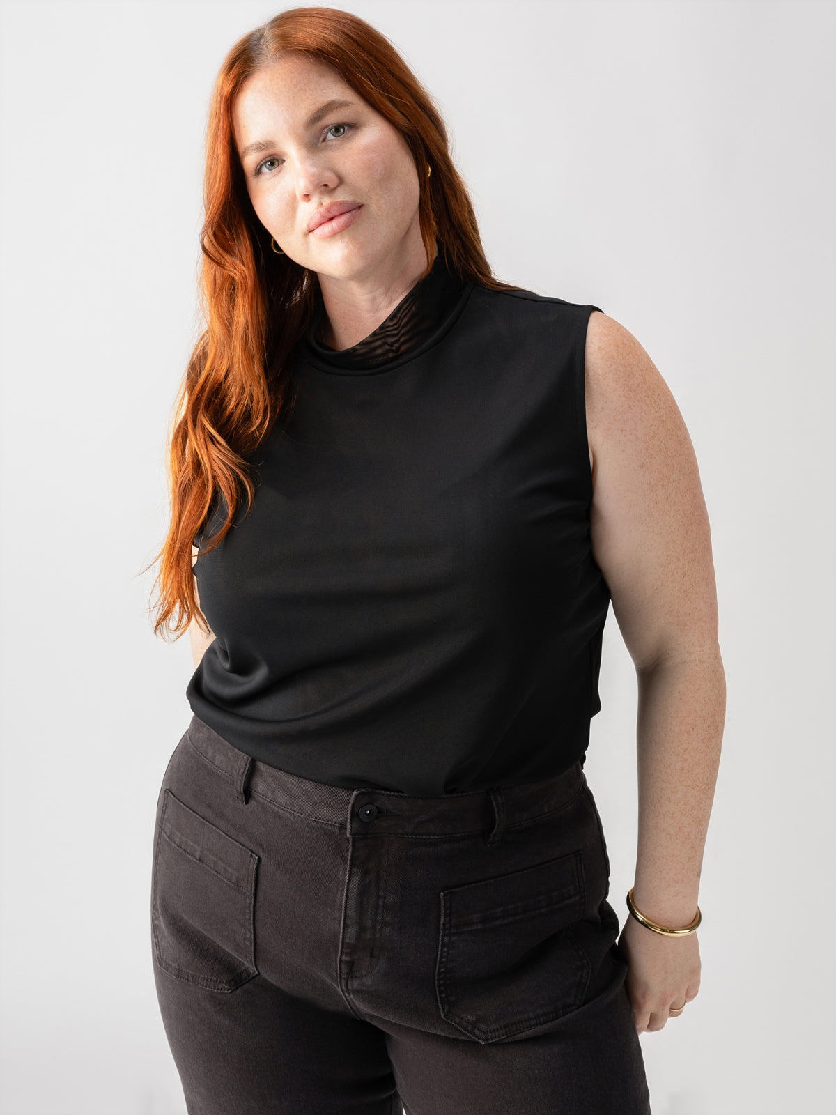 A person with long red hair is standing against a plain background, dressed in the Sanctuary Clothing Shirred Neck Mesh Tank Black from the Inclusive Collection and dark pants. Their right hand is resting on their hip and they are looking at the camera with a neutral expression.