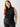 A person with long red hair is standing against a plain background, dressed in the Sanctuary Clothing Shirred Neck Mesh Tank Black from the Inclusive Collection and dark pants. Their right hand is resting on their hip and they are looking at the camera with a neutral expression.