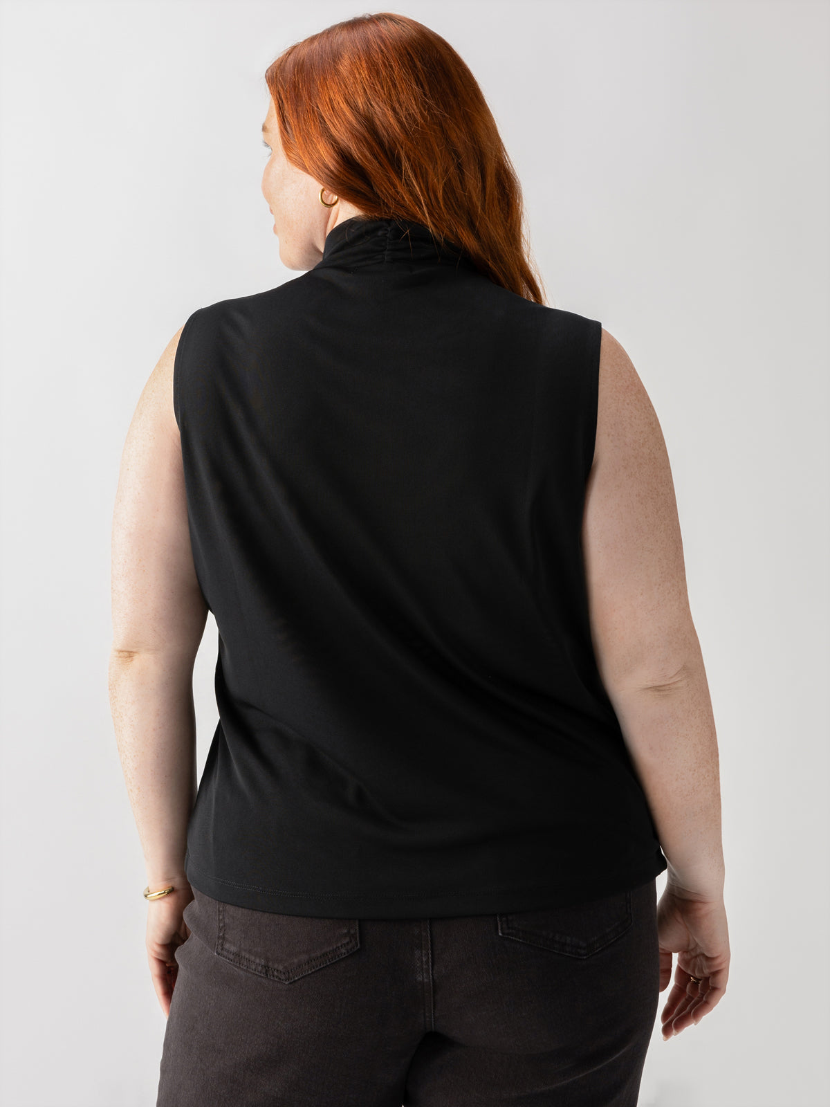 A person with long auburn hair is shown from behind, wearing Sanctuary Clothing's Shirred Neck Mesh Tank Black Inclusive Collection top and dark jeans. The background is plain and light-colored, highlighting the subject’s attire and hair.