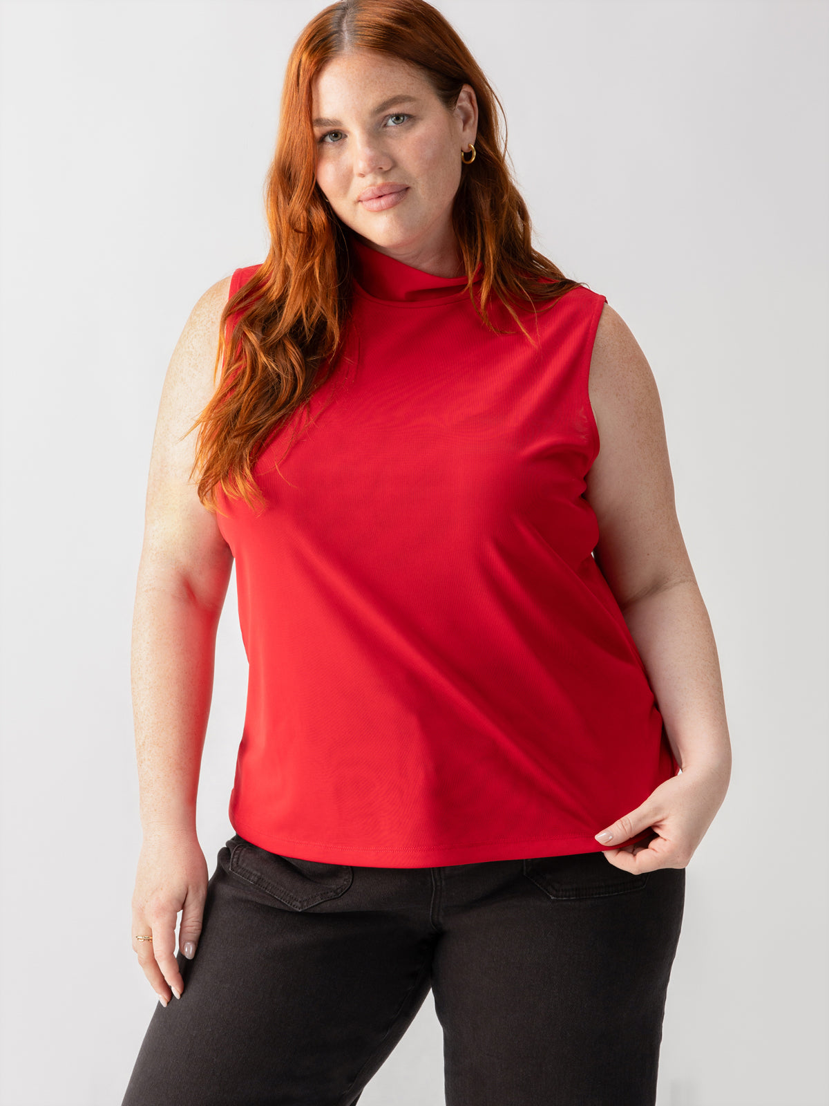 A woman with long red hair is wearing the Shirred Neck Mesh Tank in Cherry Red from Sanctuary Clothing's Inclusive Collection, paired with black jeans. She stands against a plain white background, holding the hem of her top with one hand and looking directly at the camera with a slight smile.