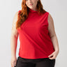 A woman with long red hair is wearing the Shirred Neck Mesh Tank in Cherry Red from Sanctuary Clothing's Inclusive Collection, paired with black jeans. She stands against a plain white background, holding the hem of her top with one hand and looking directly at the camera with a slight smile.