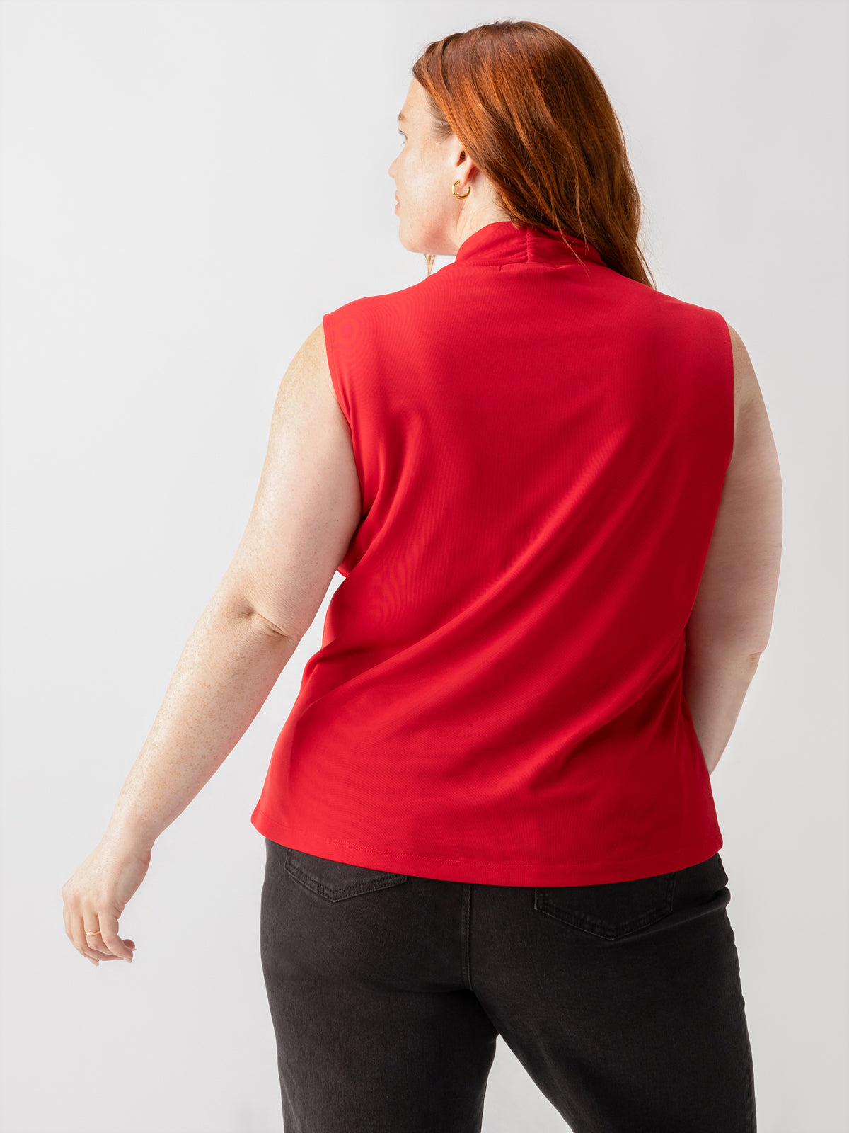 A person with long red hair is standing with their back to the camera, wearing the Shirred Neck Mesh Tank Cherry Red from Sanctuary Clothing's Inclusive Collection and a pair of black pants. The background is a plain, light-colored wall.