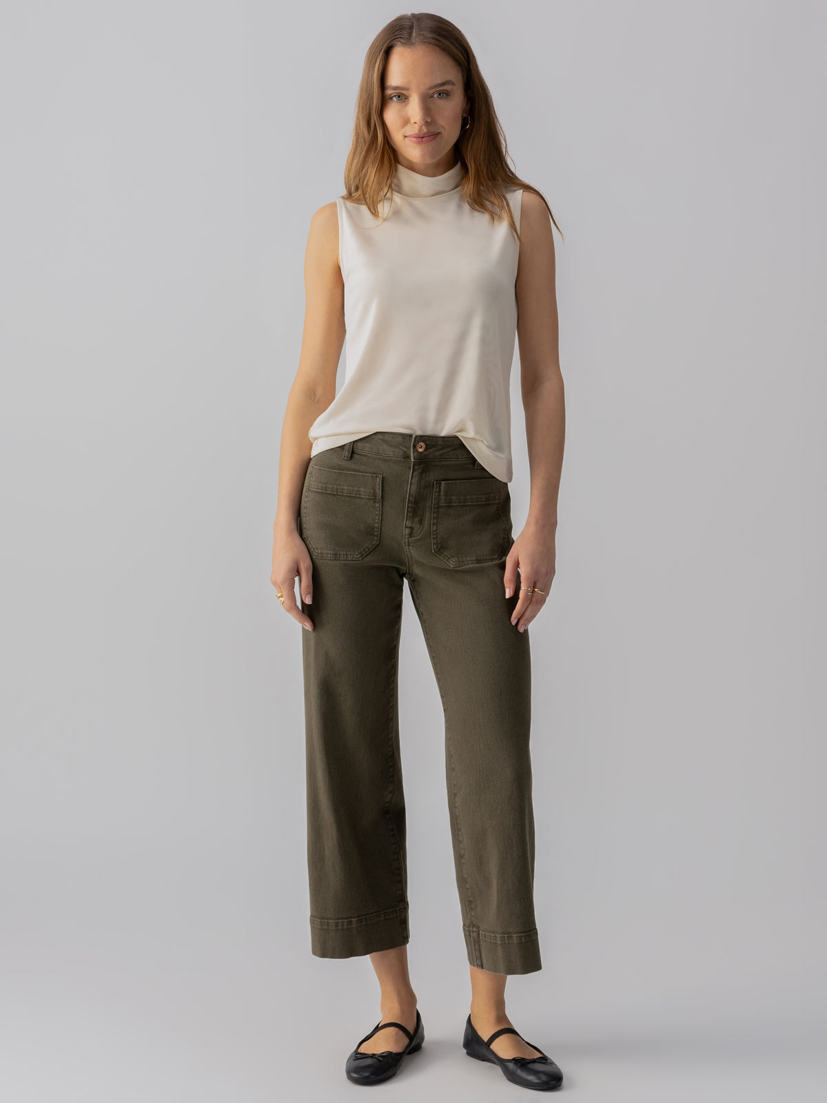 A woman stands against a plain backdrop, wearing the Shirred Neck Mesh Tank Birch from Sanctuary Clothing and high-waisted, olive green cropped pants. She completes her outfit with black ballet flats. Her hair is down and she gazes directly at the camera with a neutral expression.