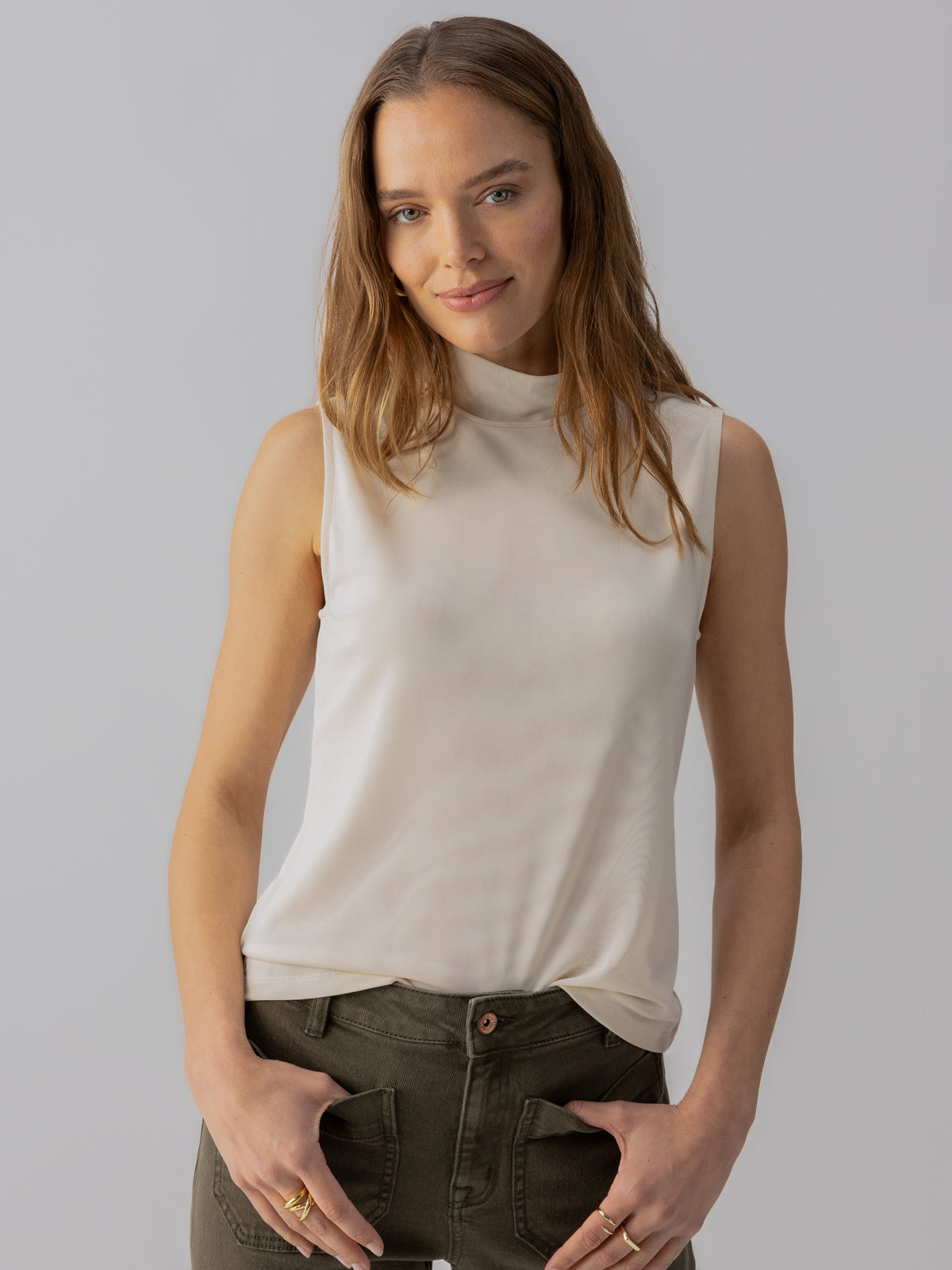 A woman with long, light brown hair and a slight smile is wearing the Shirred Neck Mesh Tank Birch from Sanctuary Clothing paired with green pants. She has her hands in her pockets and stands against a light gray background.