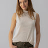 A woman with long, light brown hair and a slight smile is wearing the Shirred Neck Mesh Tank Birch from Sanctuary Clothing paired with green pants. She has her hands in her pockets and stands against a light gray background.