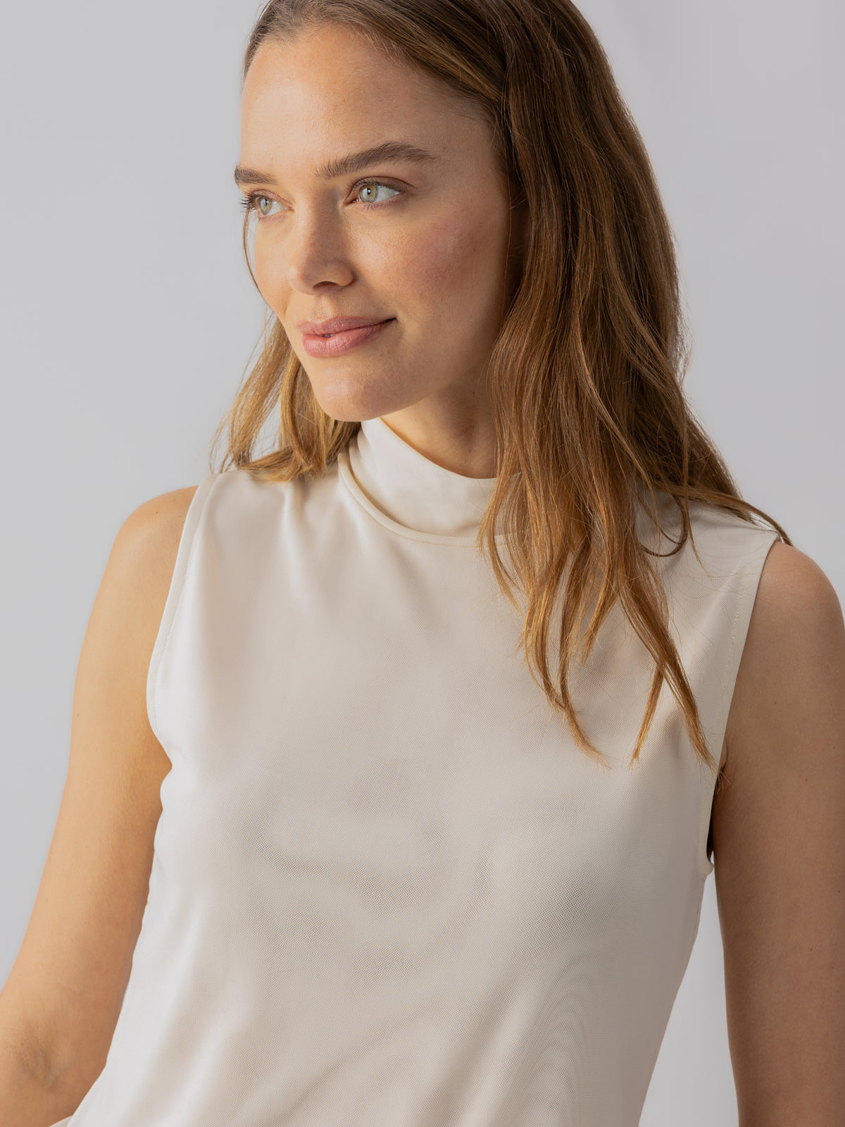 A woman with long light brown hair is smiling and looking slightly to the side. She is wearing the sanctuary clothing's Shirred Neck Mesh Tank in Birch, a sleeveless, high-neck ivory top. The background is simple and light-colored, keeping the focus on her.