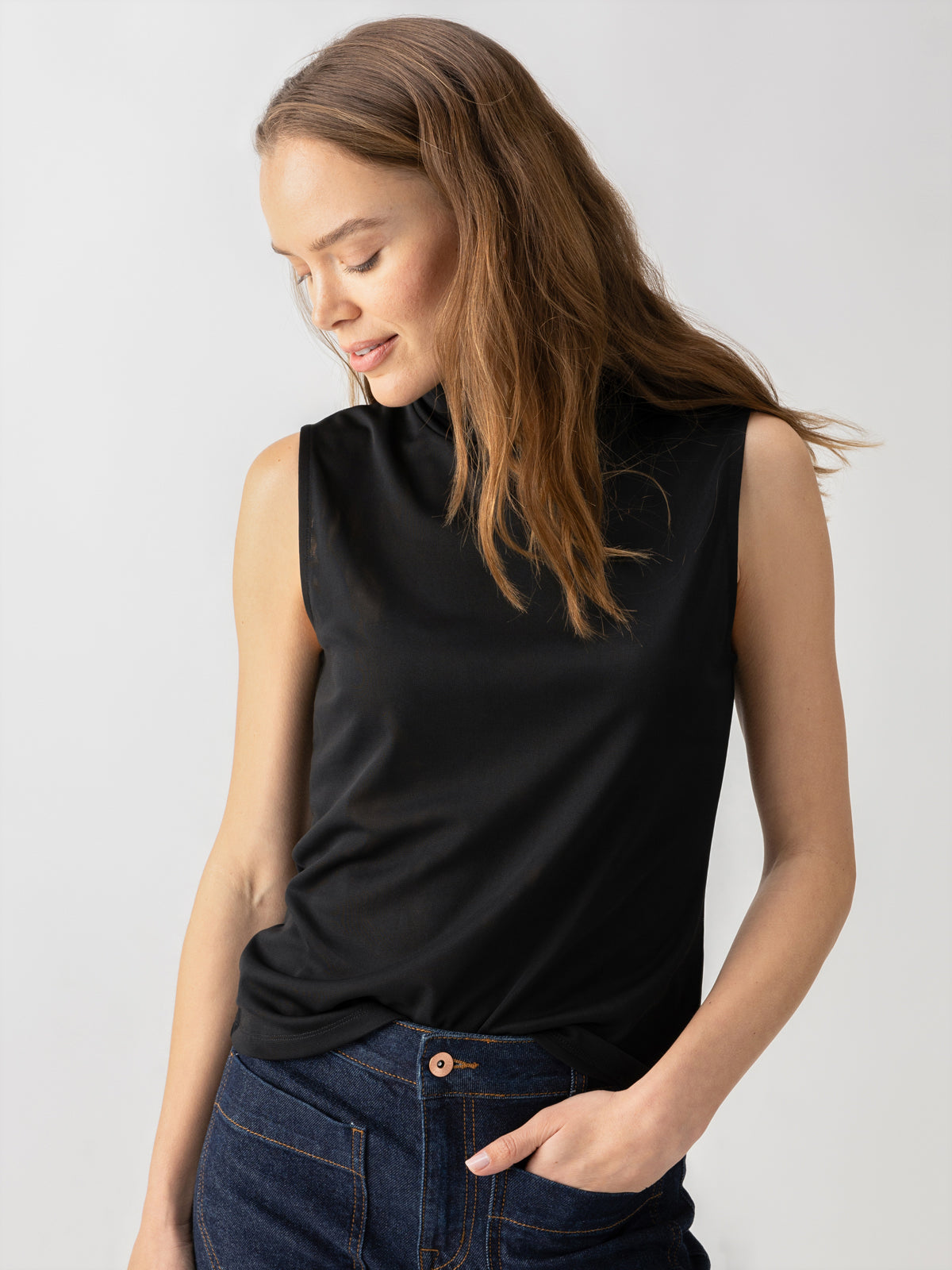 A woman with long brown hair is wearing the Shirred Neck Mesh Tank in black by Sanctuary Clothing and dark blue jeans. She is looking down and slightly smiling, with her left hand in her pocket. The background is plain and light-colored.