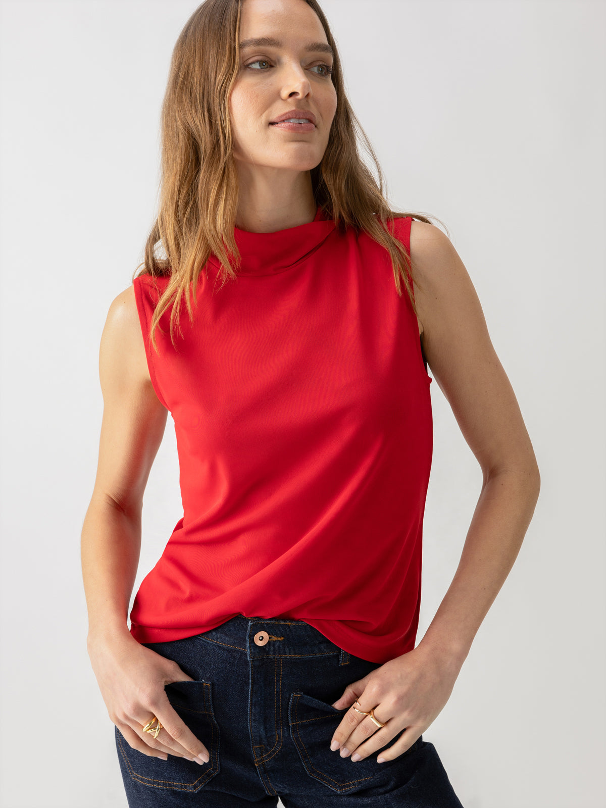 A person with long hair is wearing Sanctuary Clothing's Shirred Neck Mesh Tank in Cherry Red and dark jeans, with hands in the pockets. The individual is looking slightly to the side and has a relaxed expression against a plain light grey background.