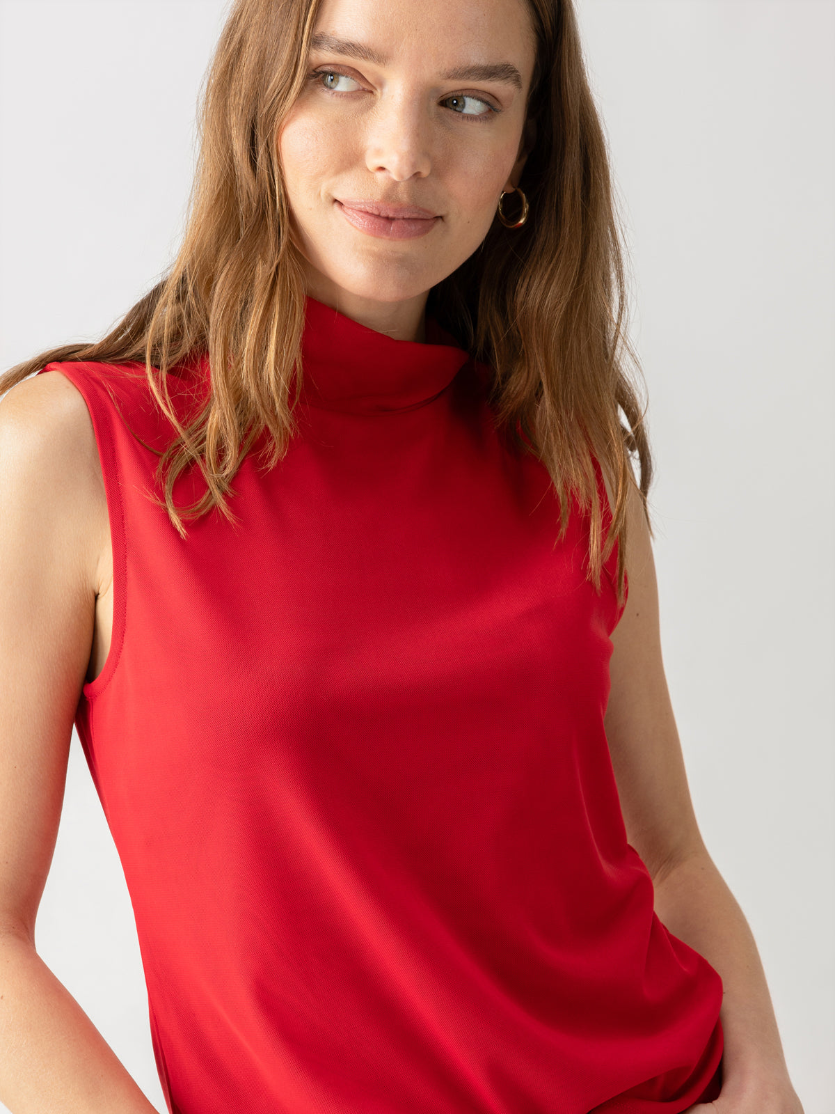 A woman with long, light brown hair is wearing a Shirred Neck Mesh Tank in Cherry Red by Sanctuary Clothing. She is smiling slightly and looking to her right, with a neutral grey background behind her. She is also wearing small hoop earrings.