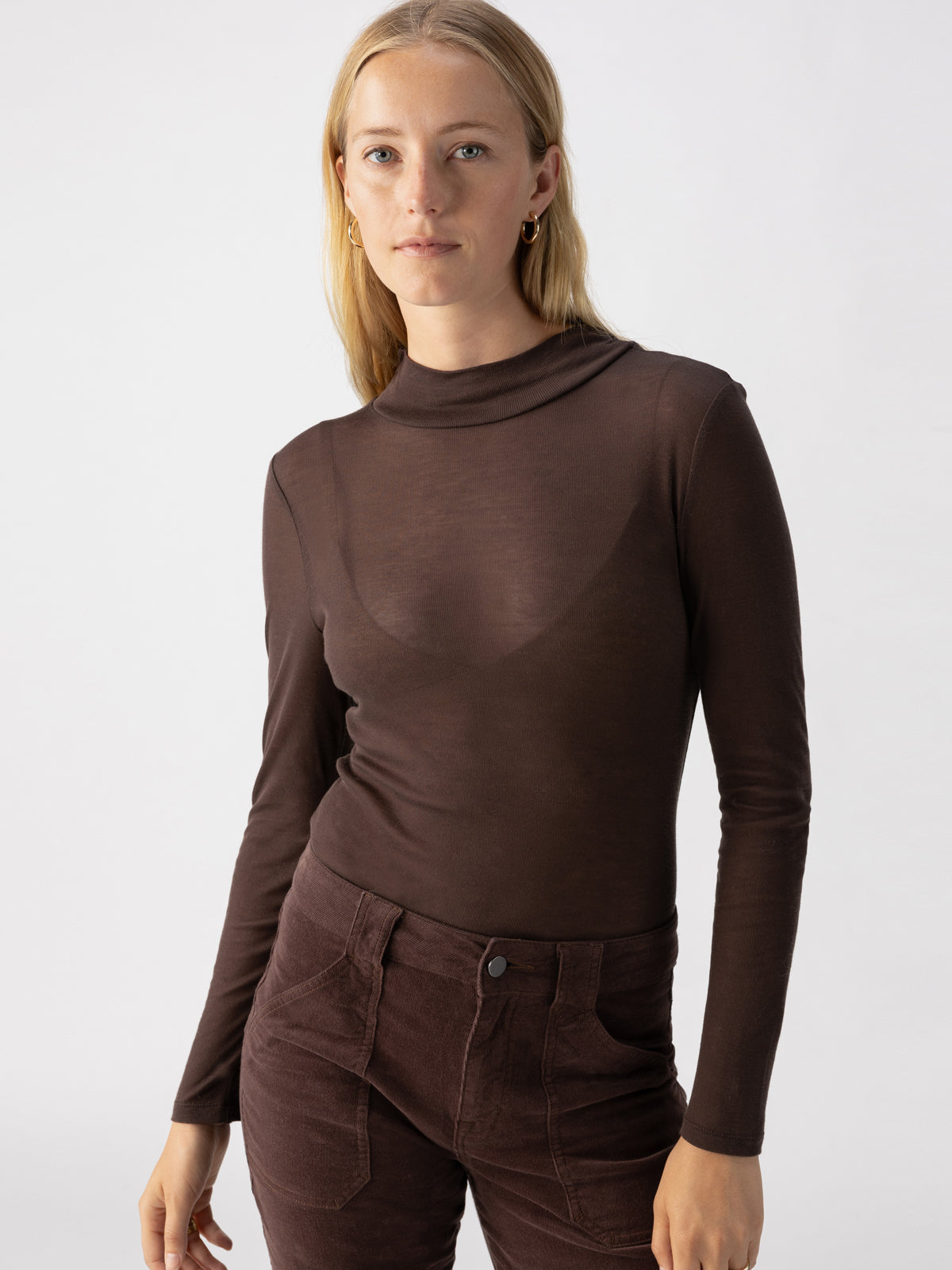 A woman with shoulder-length blonde hair is wearing the Semi Sheer Long Sleeve Mock Coffee by Sanctuary Clothing and high-waisted brown pants. She is standing against a plain, light gray background and looking directly into the camera with a neutral expression.