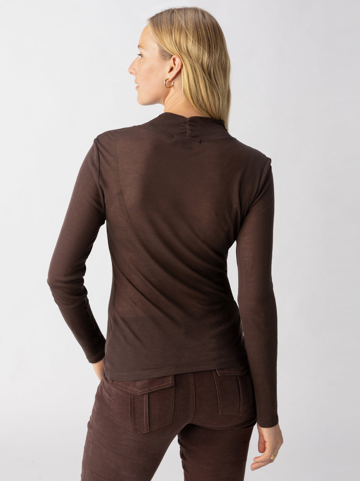 A person with long blonde hair wearing The Semi Sheer Long Sleeve Mock Coffee top from Sanctuary Clothing and brown pants is standing with their back to the camera, looking over their left shoulder against a plain white background.