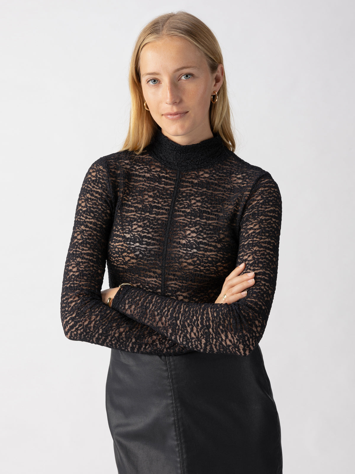 A woman with long blonde hair is standing against a plain background, wearing the Long Sleeve Textured Mock Black top by Sanctuary Clothing and a black skirt. Her arms are crossed, and she has a slight smile on her face. She is also accessorizing with small hoop earrings.