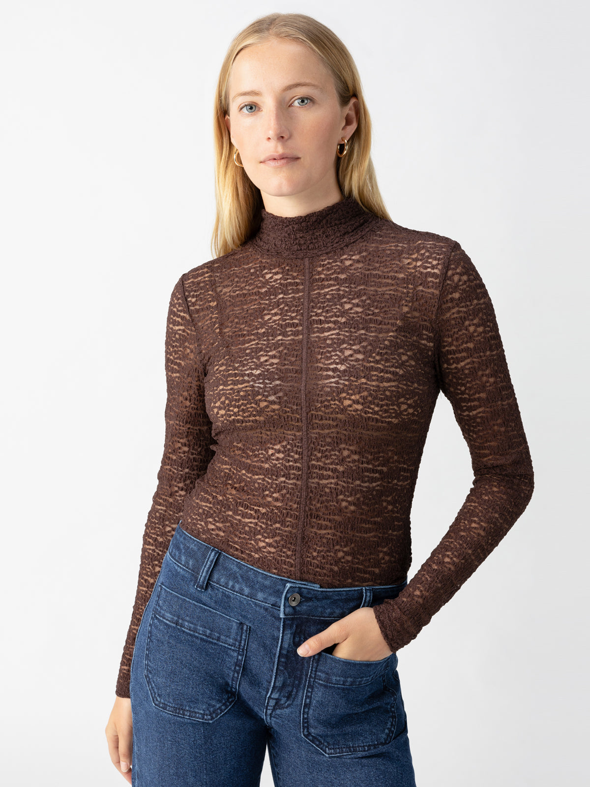 A woman is standing against a plain background, wearing the Long Sleeve Textured Mock Brown Sugar top from Sanctuary Clothing and high-waisted blue jeans. She has long blonde hair and is looking directly at the camera with a neutral expression, with one hand in her pocket.