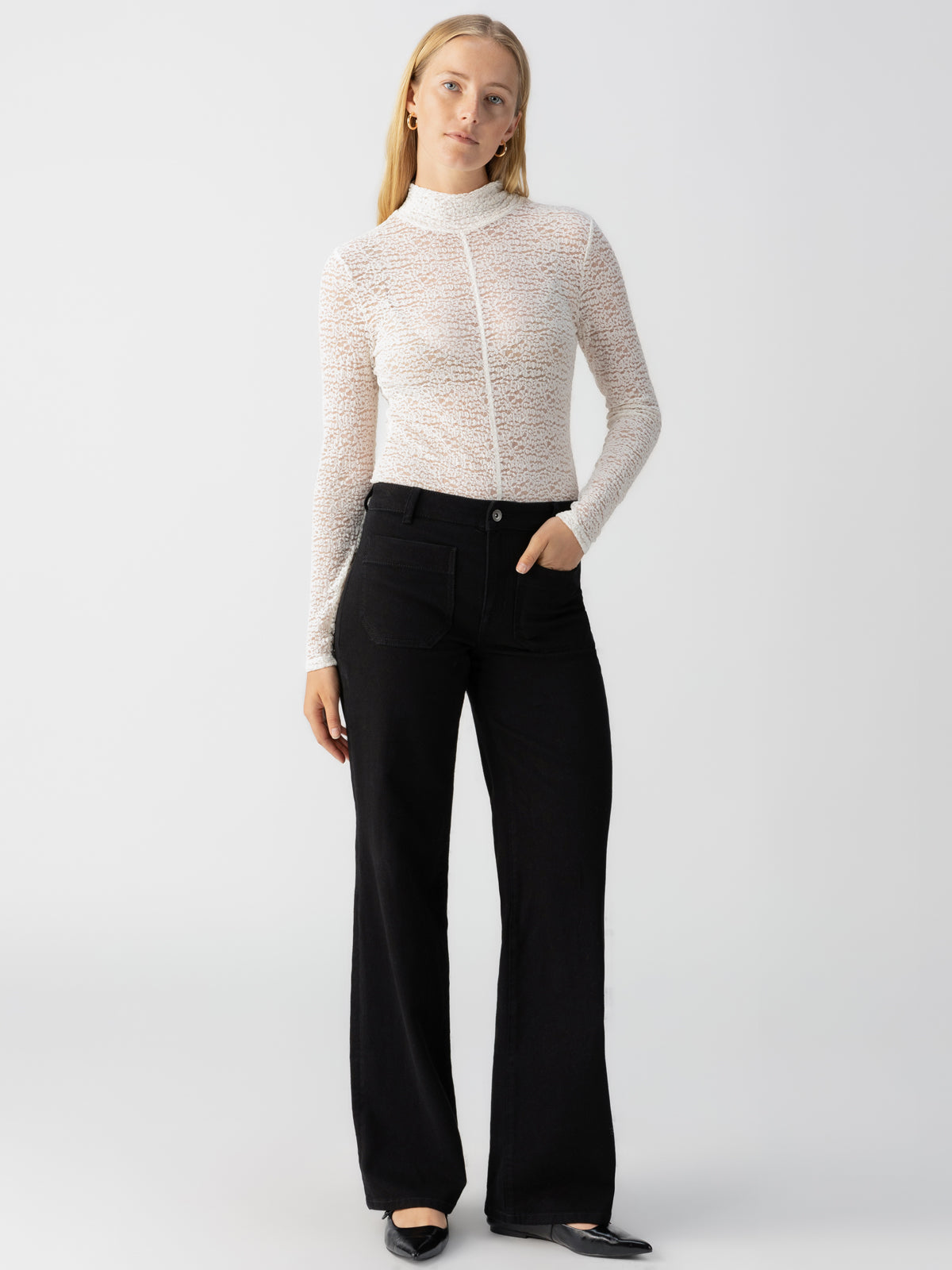 A person stands against a plain background wearing Sanctuary Clothing's Long Sleeve Textured Mock Chalk top paired with high-waisted black wide-leg pants. They have long blonde hair, are sporting black flats, and display a relaxed facial expression and posture.