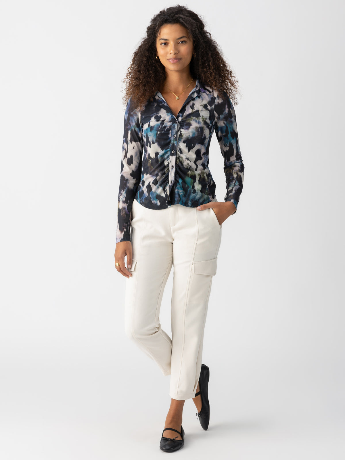 A woman with curly hair is wearing the Sanctuary Clothing Dreamgirl Mesh Button Up Midnight Storm, a long-sleeved, patterned blouse in shades of blue, black, and white. She has on white, straight-leg pants with side pockets and black flat shoes. She stands against a plain white background, with one hand in her pocket.