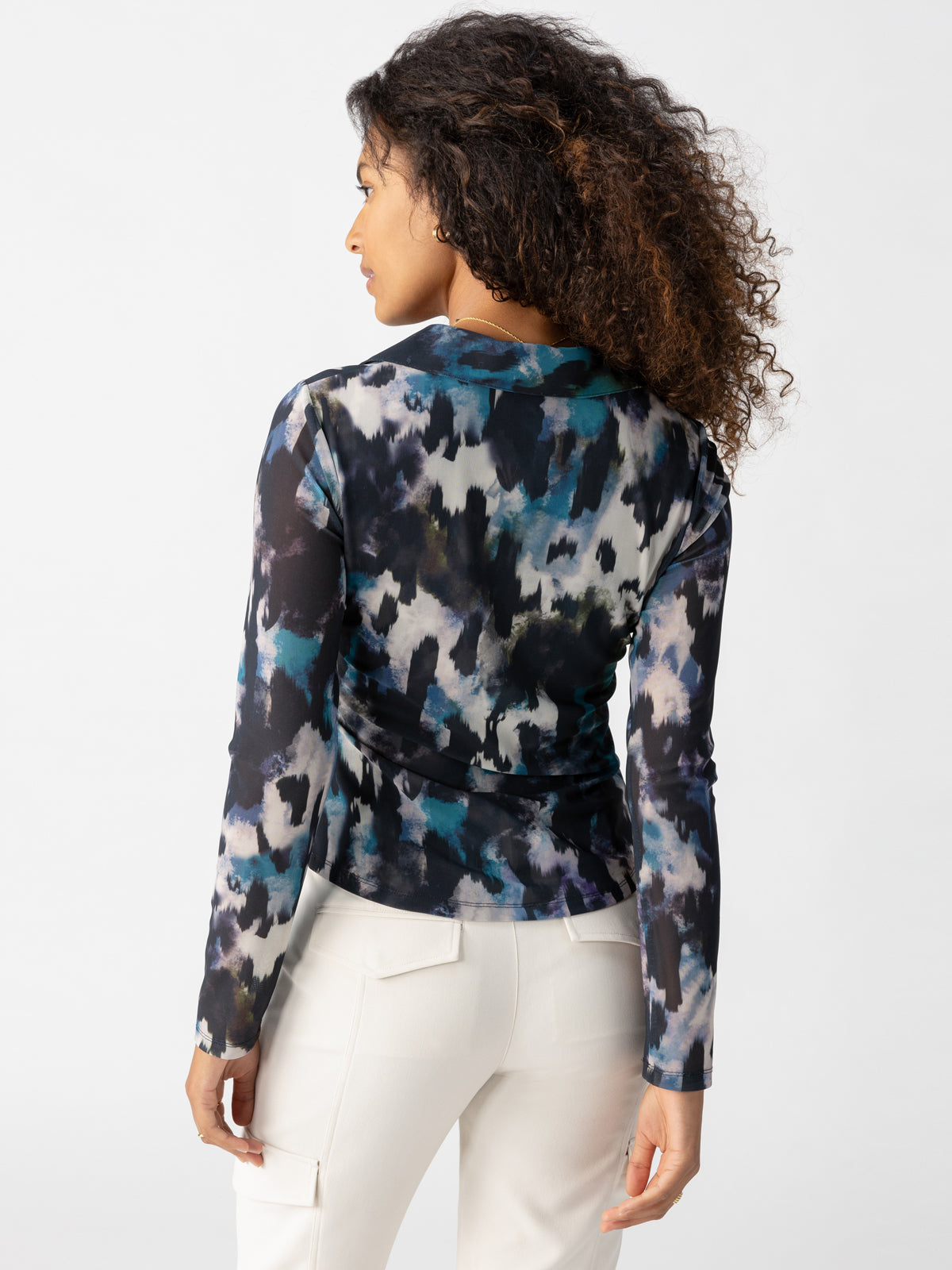 A woman with curly hair is shown from the back, wearing the Dreamgirl Mesh Button Up Midnight Storm by Sanctuary Clothing. The top features an abstract, multicolored pattern in shades of blue, black, and white. She has paired it with high-waisted white pants. The background is a plain light gray.